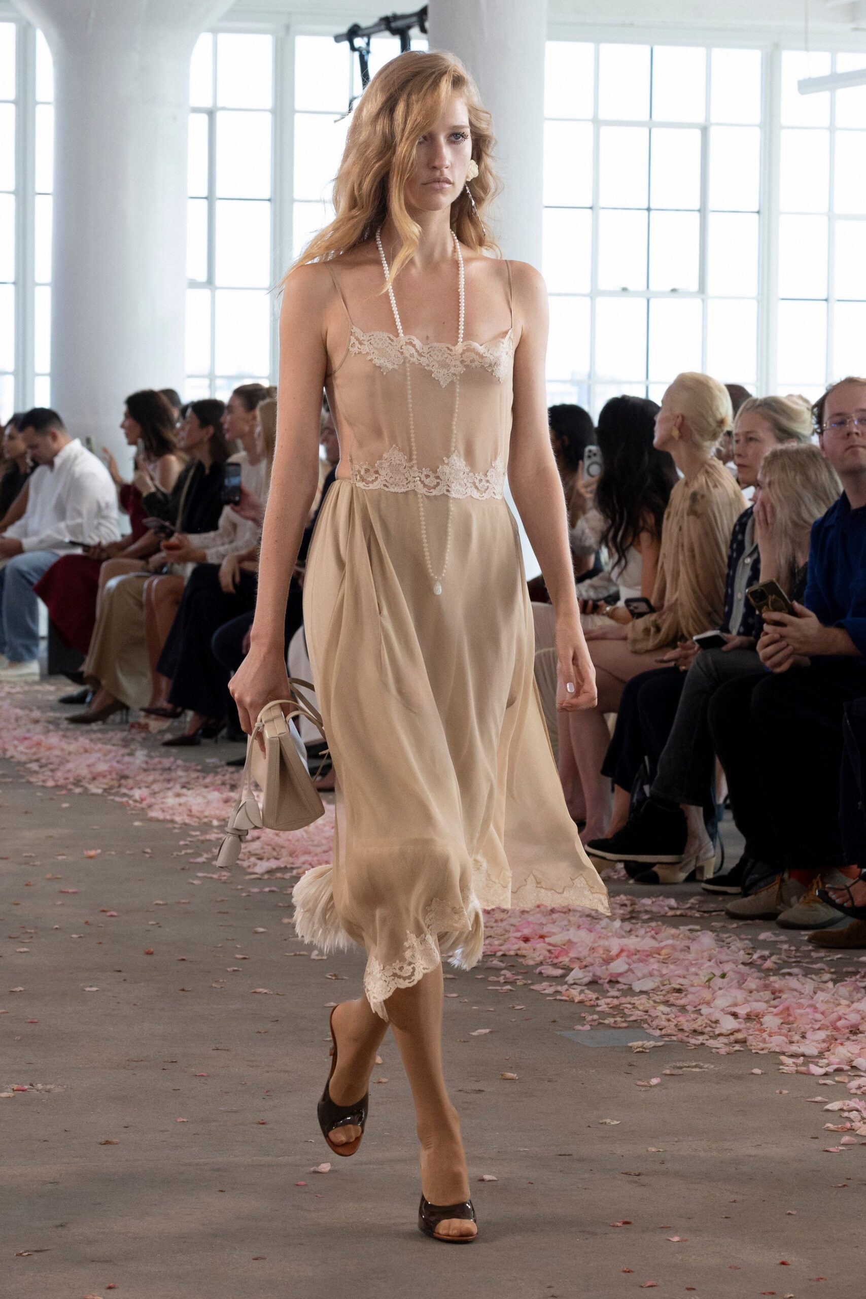 Champagne satin slip dress with scalloped lace at bust, waist, and uneven hem. Long pearl strand ending in a teardrop pendant. Cream top-handle bag with a knotted strap tail. Dark patent peep-toe mules. Fluffy white cuffs circling both ankles. Walking home from a courthouse wedding with petals stuck to the pavement.