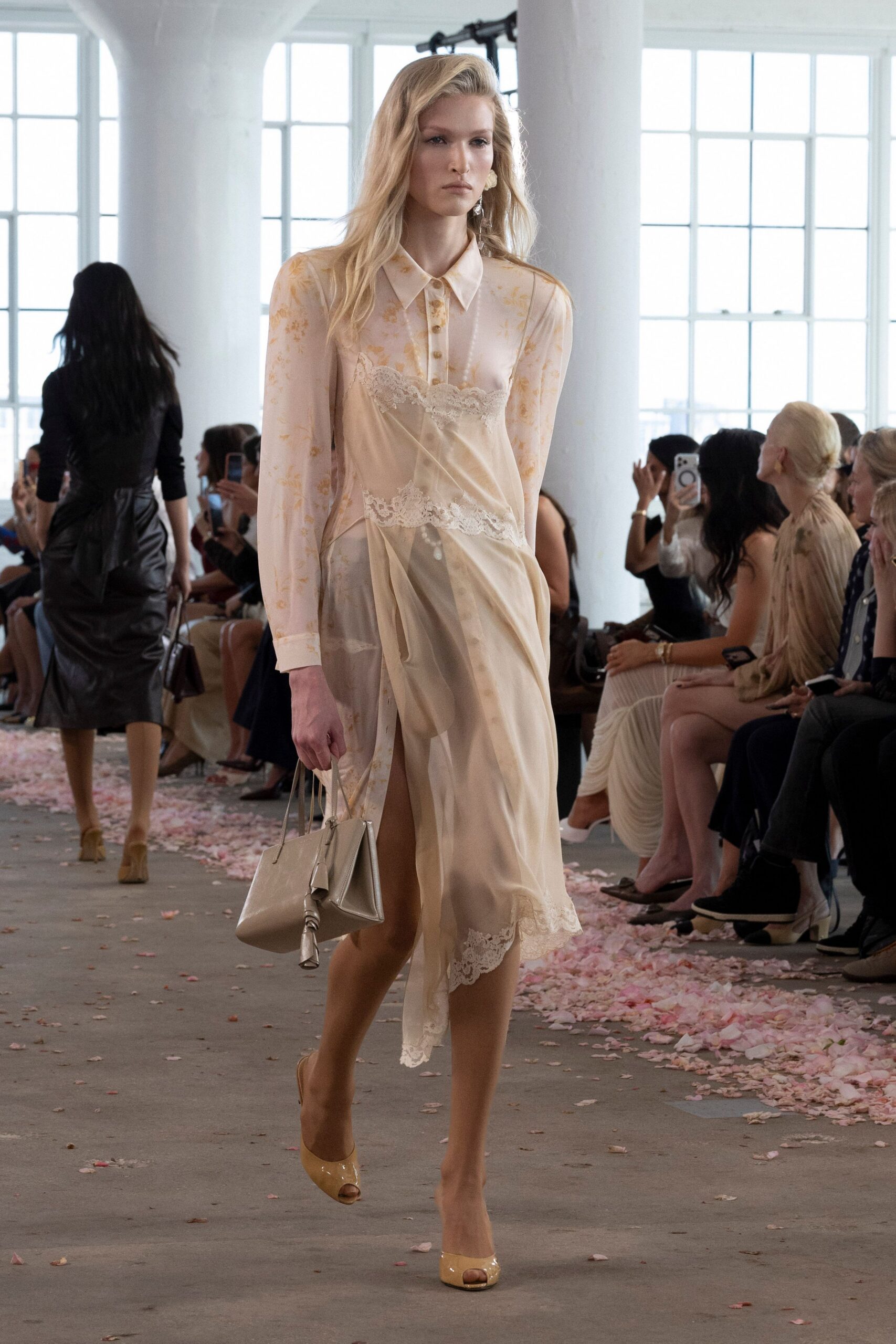 Sheer blush blouse scattered with faint apricot florals and pearl buttons. Bias-cut champagne slip, lace bands interrupting the draped front slit. Glossy taupe tote with a tasselled zip pull swinging low. Cream patent peep-toes. Hotel brunch after the wedding, petals still stuck to the pavement outside.