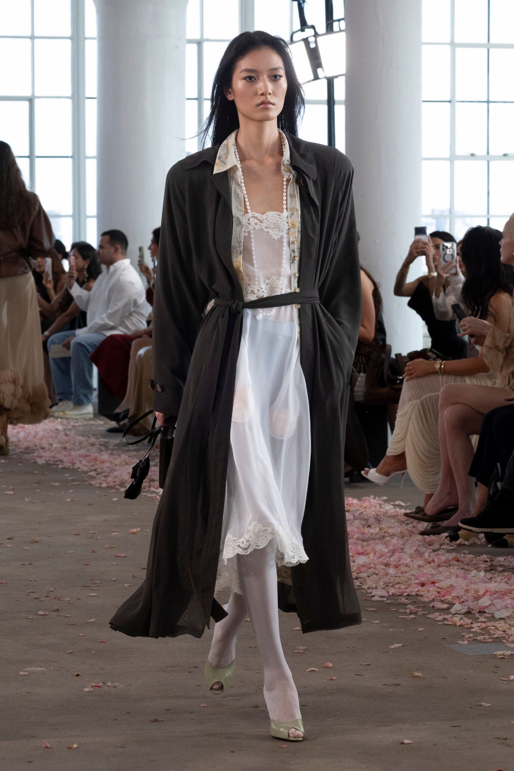 Charcoal sheer coat, wrapped shut with a narrow self belt. White satin slip with scalloped lace at bust and hem. Long pearl strand hanging straight past the waist. Opaque white tights. Pistachio peep-toe mules. For crossing a hotel corridor at dawn, bouquet petals stuck to the carpet.
