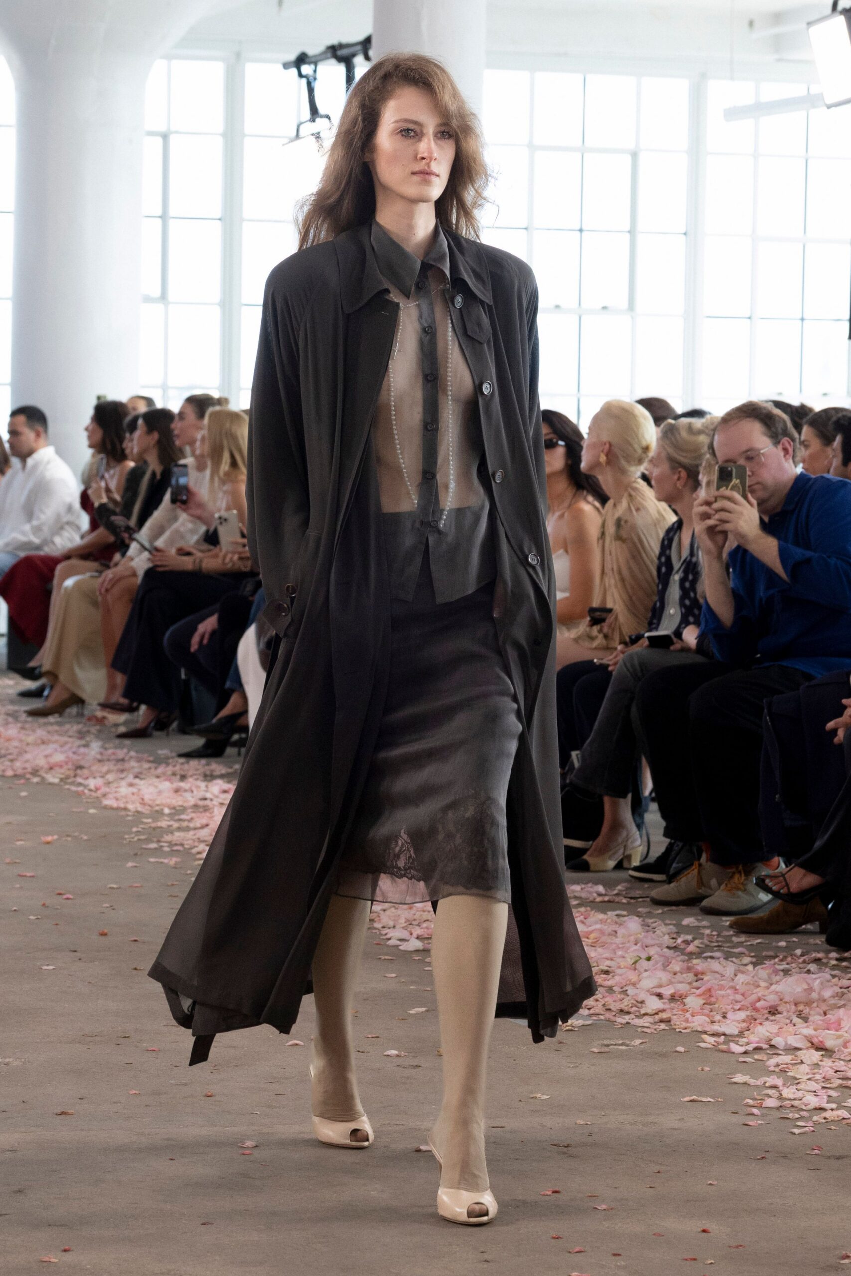 Long charcoal coat in fluid matte weave, one cuff tab hanging loose. Sheer taupe shirt with smoke-grey placket and tiny black buttons. Charcoal satin slip skirt with a shadowy lace bloom at the hem. Barely beige sheer tights. Cream peep-toe heels. Walking through a conservatory after the wedding flowers have started dropping.