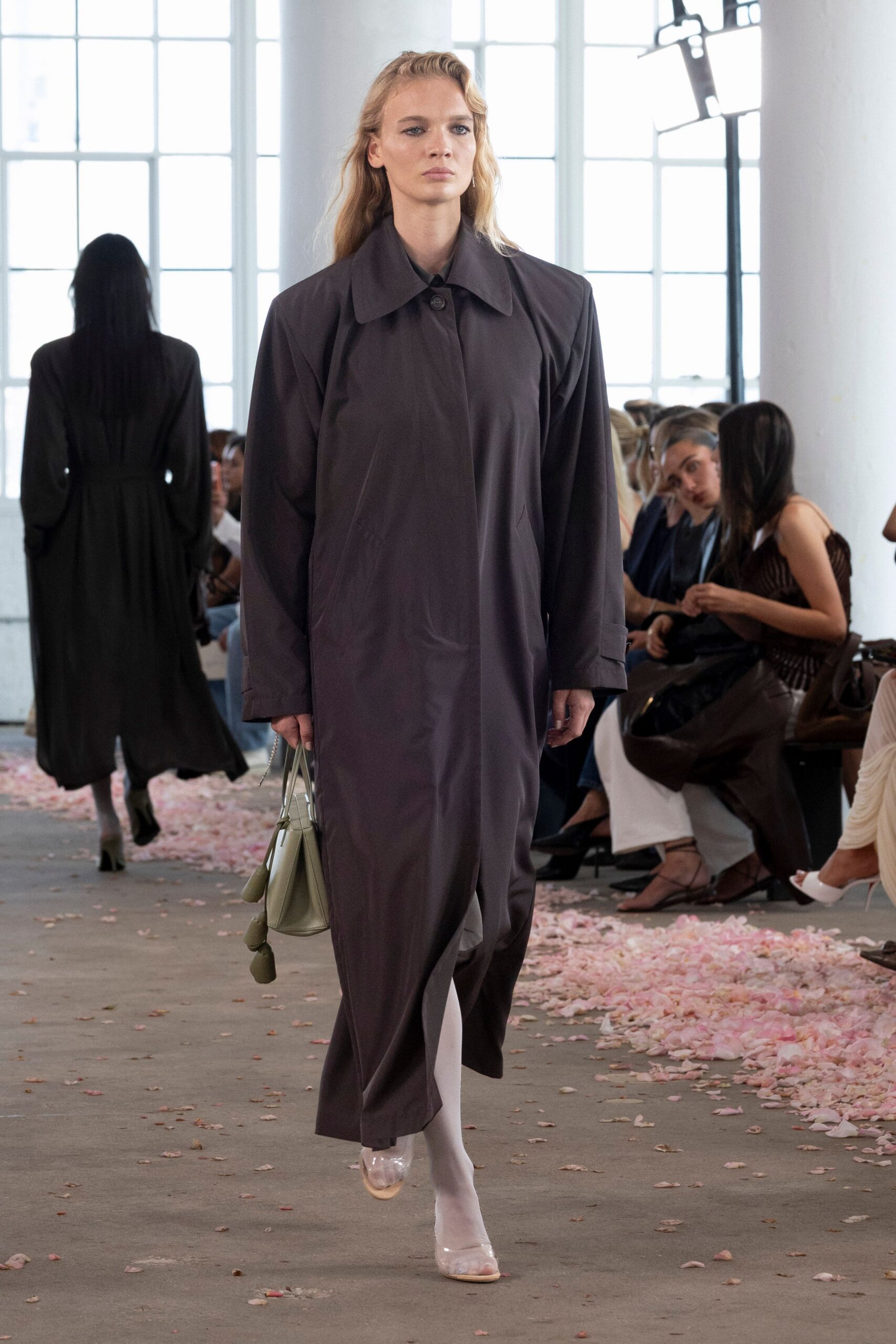 Charcoal coatdress in crisp twill, one dark button under a wide collar. Matching straight trousers, a pale inner panel flashing at the stride. Opaque white tights. Clear pumps with nude soles. Sage trapeze bag with two pod charms swinging low. Crossing a lobby still scattered with petals after the wedding lunch.