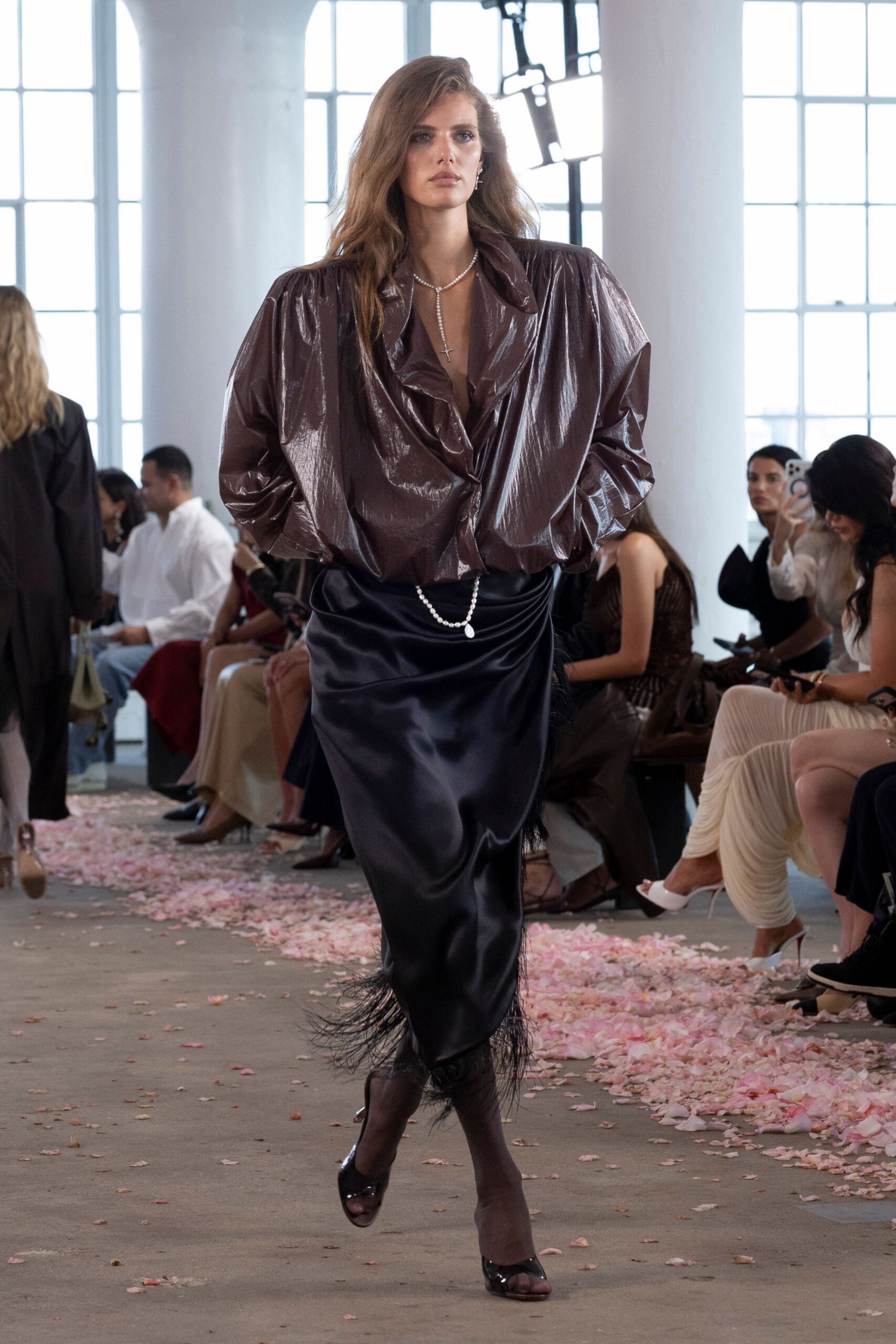 Glossy mahogany blouse with huge padded shoulders and a collapsed plunge. Pearl lariat ending in a small cross. Black satin skirt, ruched low and finished with a feathered hem. Sheer espresso tights. Patent peep-toes. Crossing a hotel ballroom after the wedding band has packed up.