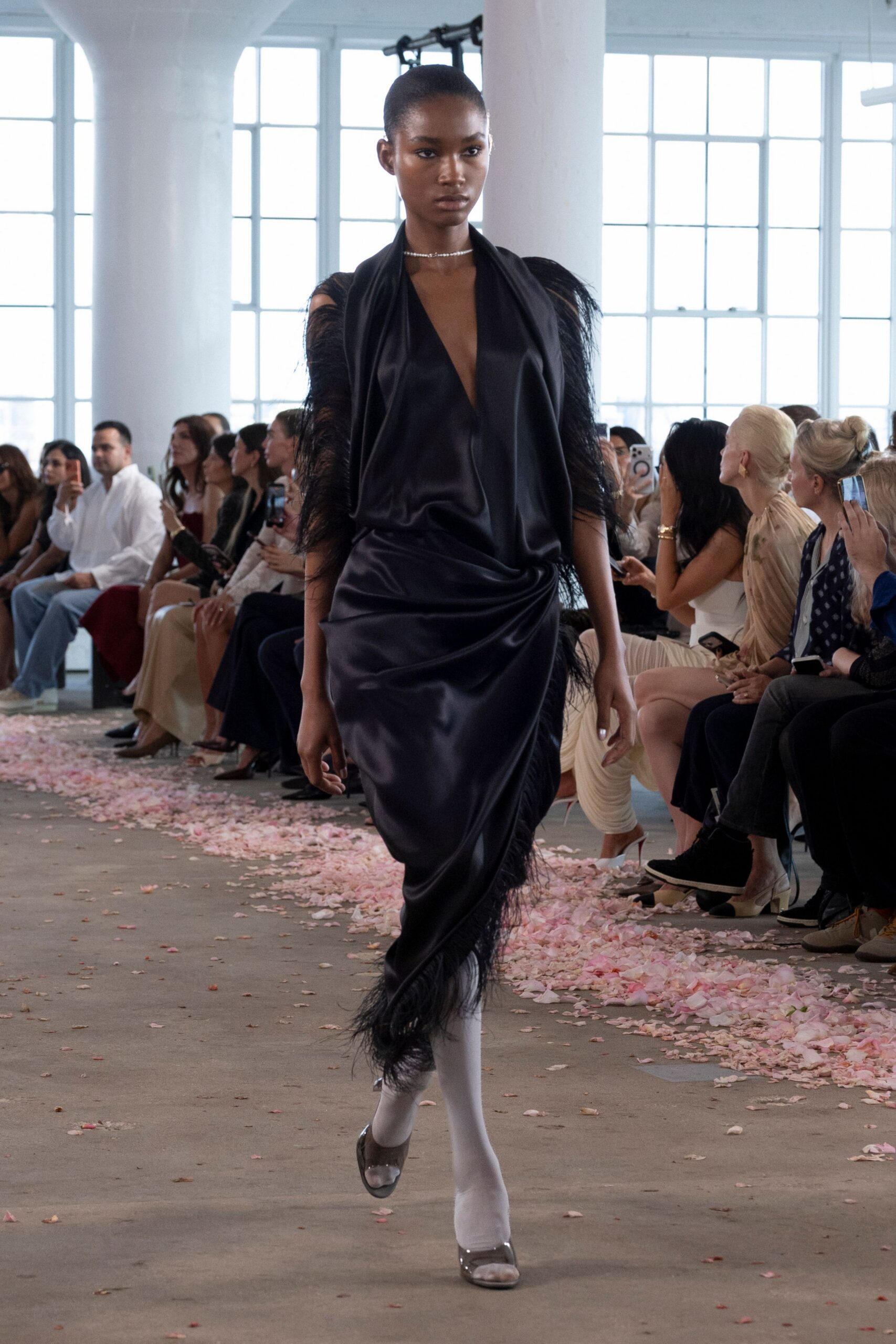 Fine crystal collar sitting close against the throat. Black liquid-satin gown with a plunging draped cowl and feathered sleeves. Opaque white tights. Smoke-clear peep-toe heels. Crossing a hotel ballroom after the flower girls have already left.