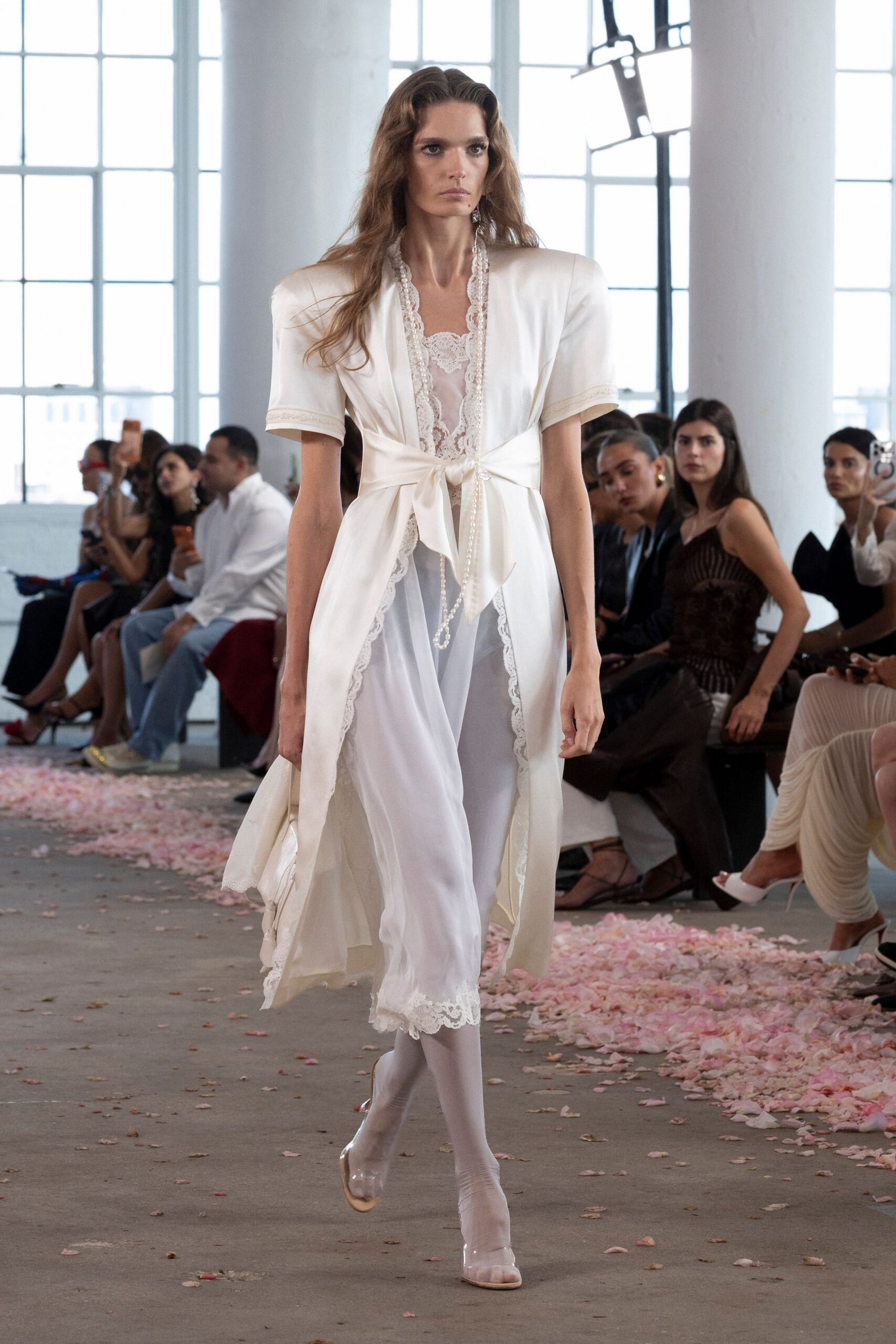 Ivory satin robe coat with pagoda shoulders and a tied waist. Sheer white slip edged in scalloped lace, hanging below the coat. Long pearl strand slipping past the knot. Milky white tights. Clear PVC peep-toe heels. Walking through crushed petals after a courthouse wedding nobody planned twice.