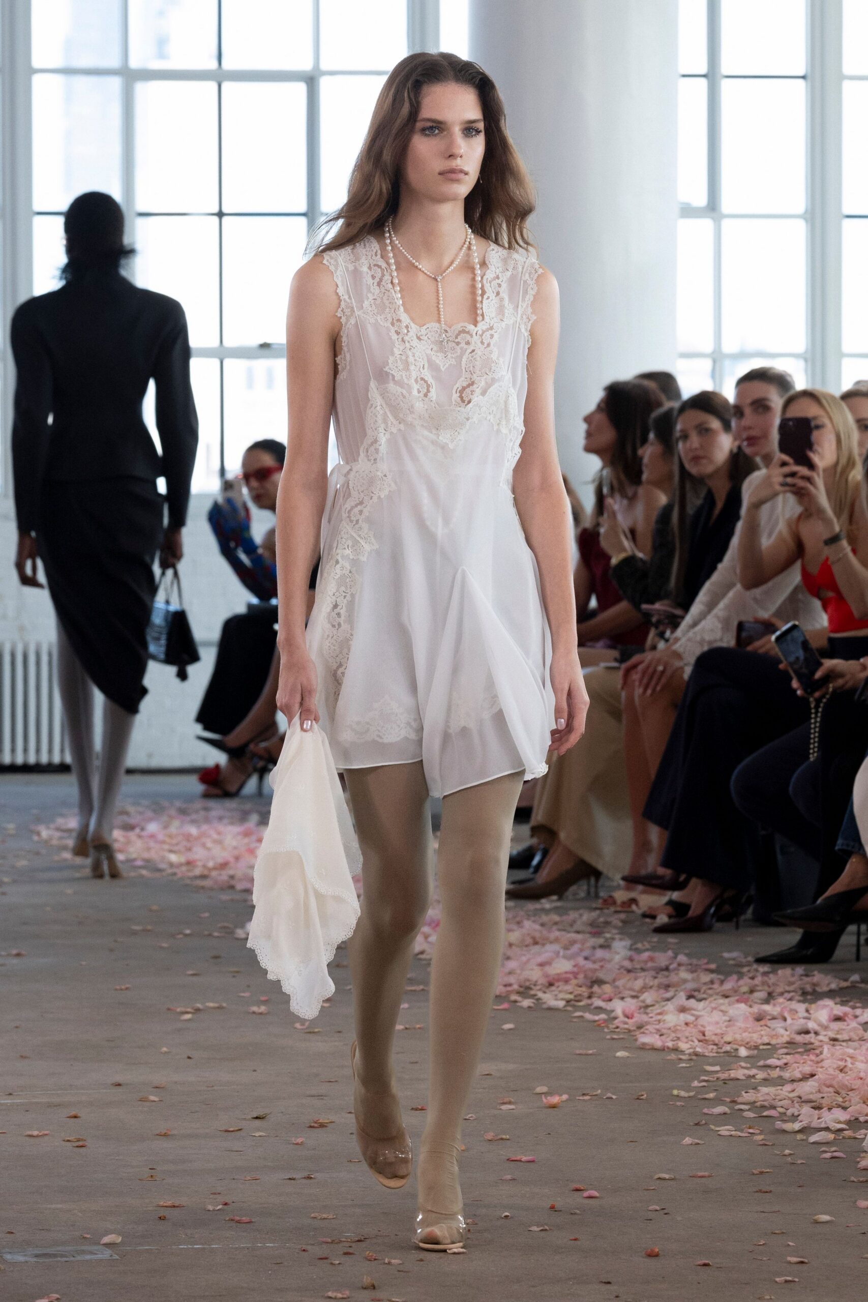Ivory chiffon slip dress paneled with scalloped lace and a dropped handkerchief hem. Double pearl strands falling into a Y at the sternum. Matching chiffon scarf, picot-edged and carried loose in one hand. Barely beige hosiery. Clear peep-toe heels. Morning after the wedding, crossing a hotel lobby still strewn with petals.