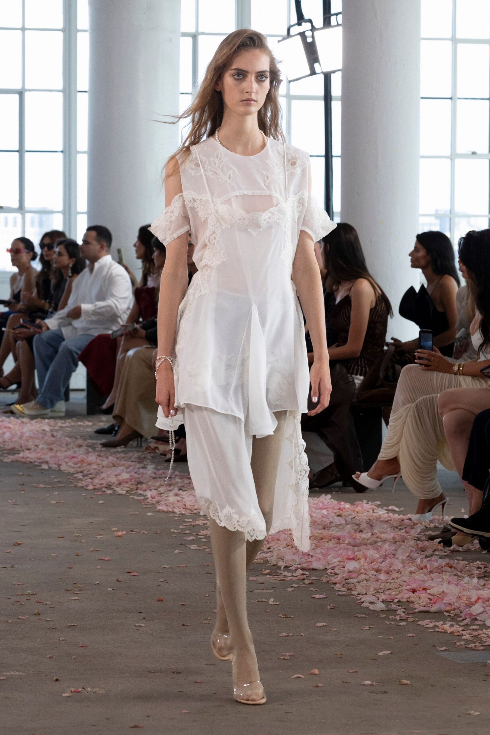 Sheer white chiffon dress with eyelash lace panels and dropped shoulder cutouts. Soft white slip peeking through the translucent bodice. Matte nude stockings. Looped pearl strands slipping from the wrist. Clear PVC pumps with a rounded toe box. Walking home from a courthouse wedding, petals stuck to the hem.