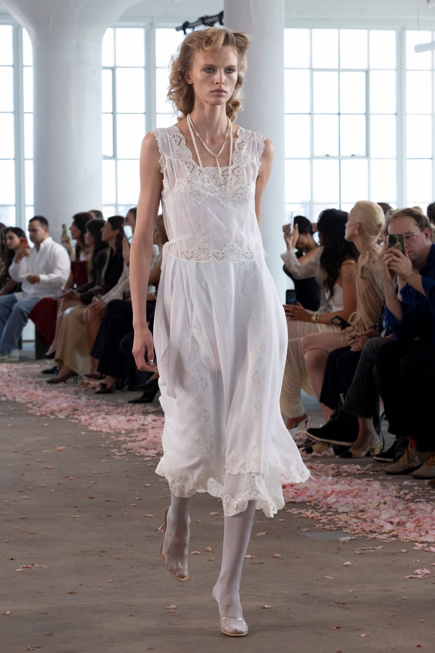 White chiffon slip dress with scalloped lace panels and a dropped waist seam. Long pearl strands doubled and trailing over the square lace neckline. Opaque white tights. Clear pumps with nude piping at the topline. Morning after the wedding, crossing a hotel ballroom still sticky with rose petals.