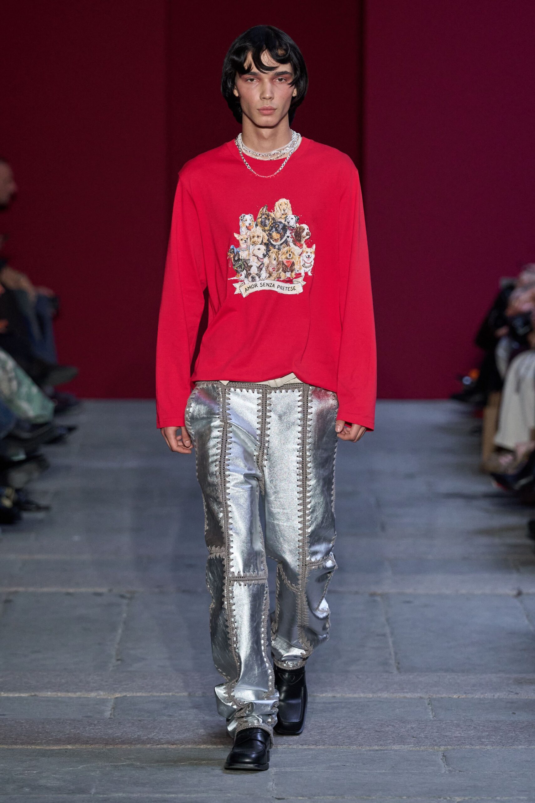 Layered silver chains sitting close at the collarbone. Cherry-red jersey tee printed with a crowded pack of dogs. Silver metallic patchwork trousers, zigzag blanket-stitch seams running vertically. Black square-toe leather shoes. Walking into a dog-show raffle still wearing last night's stage clothes.