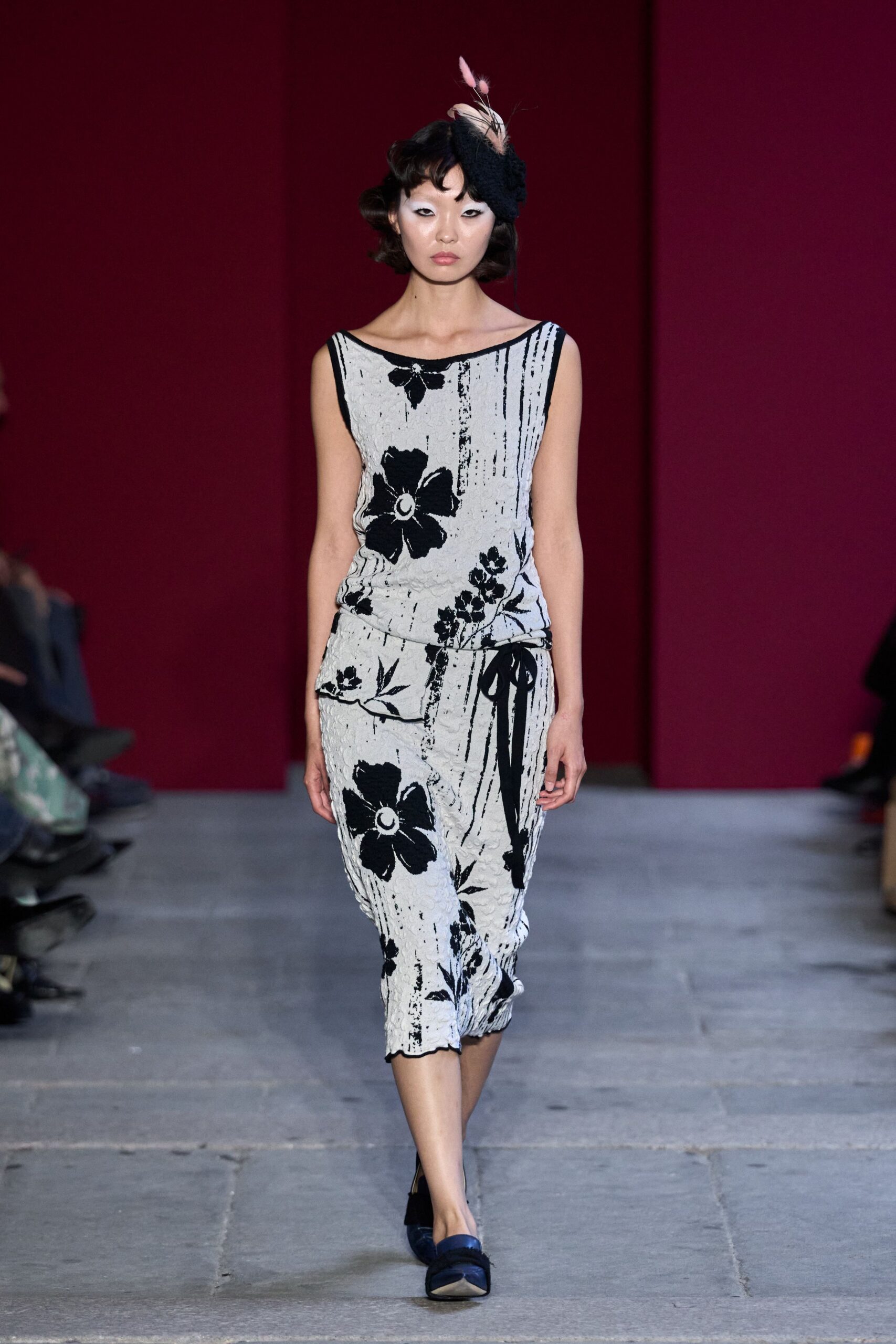 Black fuzzy pillbox with pale feathers pinned upright. Ivory crinkled knit column dress, black floral print and side sash knot. Navy satin pumps with squared toes. Hotel tea room at four, rain striping the windows.