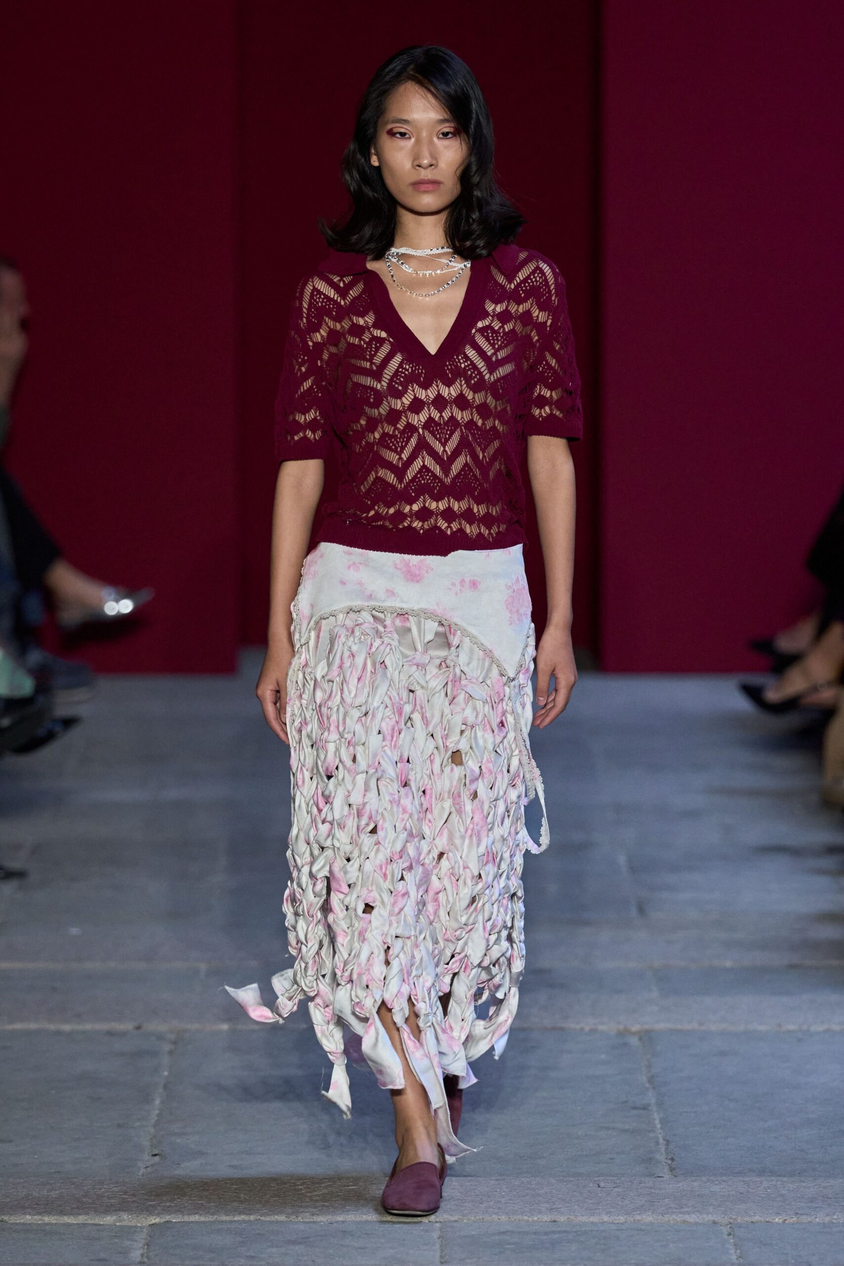 Layered silver chains with a close white crocheted strand at the throat. Mulberry openwork knit top, deep V and elbow sleeves. White skirt pieced from knotted rag strips, washed with pale pink. Mulberry suede mules. Walking through a rose garden after rain, hems already catching leaves.