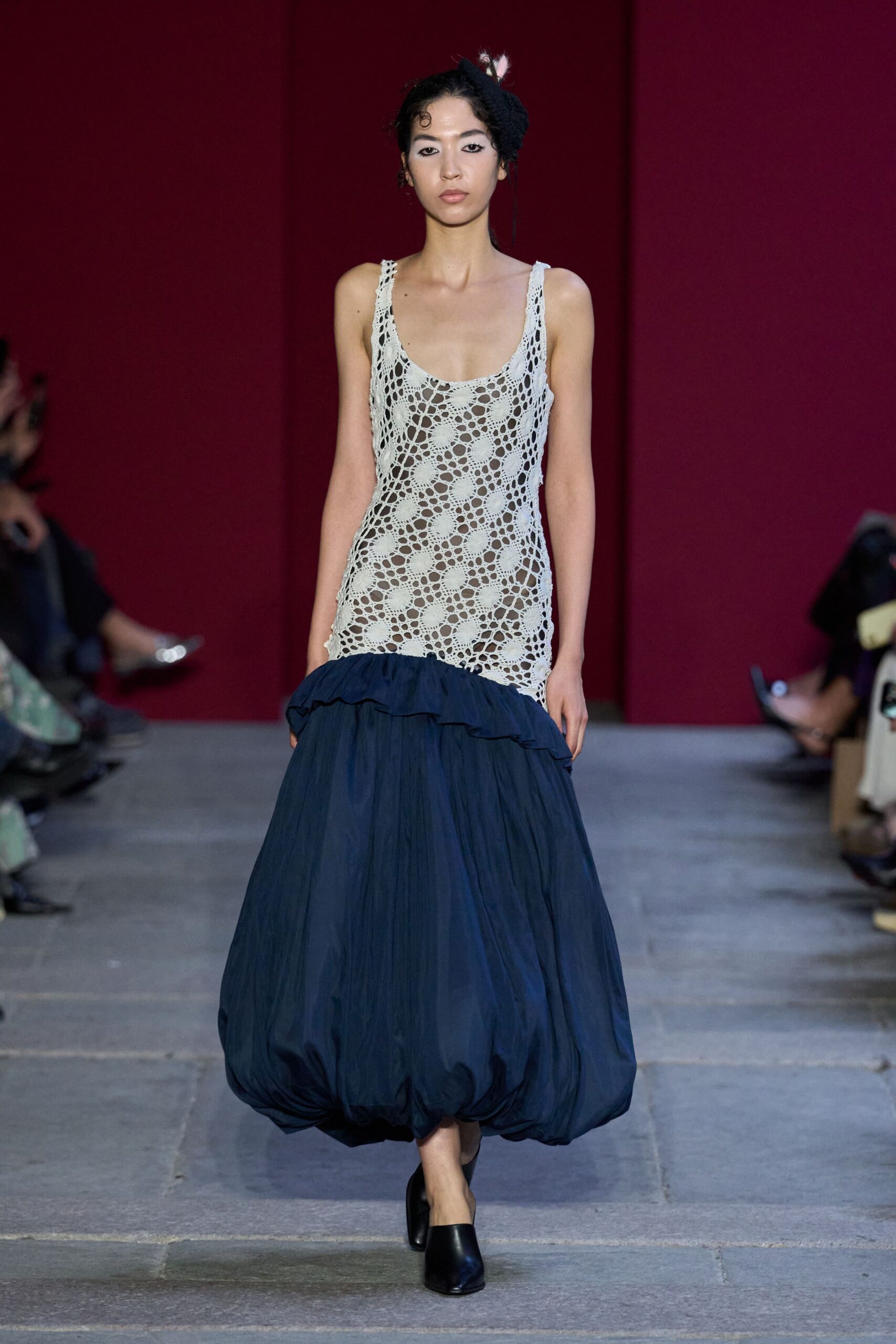 Black knit fascinator with two small white feathers poking upward. Ivory open-crochet tank dress, scooped low and sheer through the torso. Midnight poplin bubble skirt with a ruffled seam dropped at the hips. Black leather mules with a squared, closed toe. Crossing a stone courtyard after the string quartet has packed up.