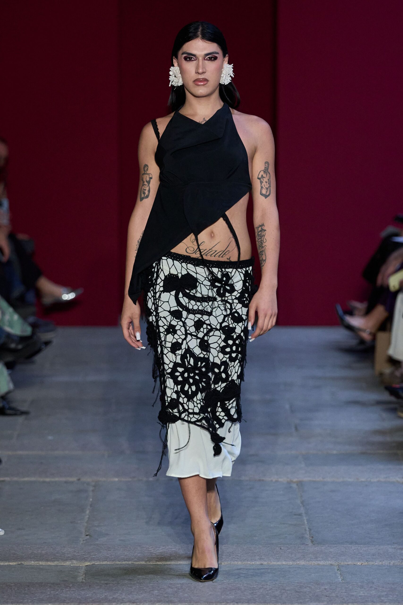 White clustered flower earrings, dense as little pompom carnations. Black halter top draped on the bias with one long pointed panel. Open black crochet flowers laid over a white slip, threads hanging loose. Black patent pumps. Walking home from a courtyard wedding after the chairs are stacked.