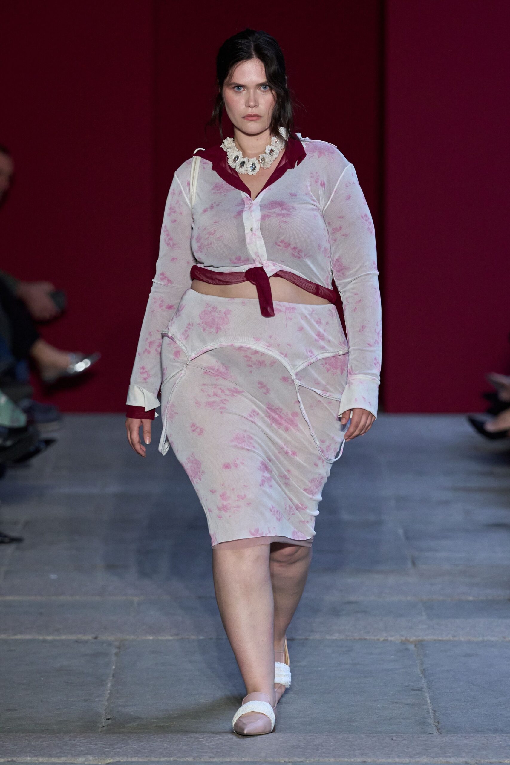 Chunky crystal bib necklace on a frayed white rosette band. Sheer white mesh blouse, pink floral print, burgundy collar and tied hem. Matching jersey skirt with curved seamed panels sitting like briefs. Blush flats with fuzzy white straps. Walking back from the hotel pool before your room key dries out.