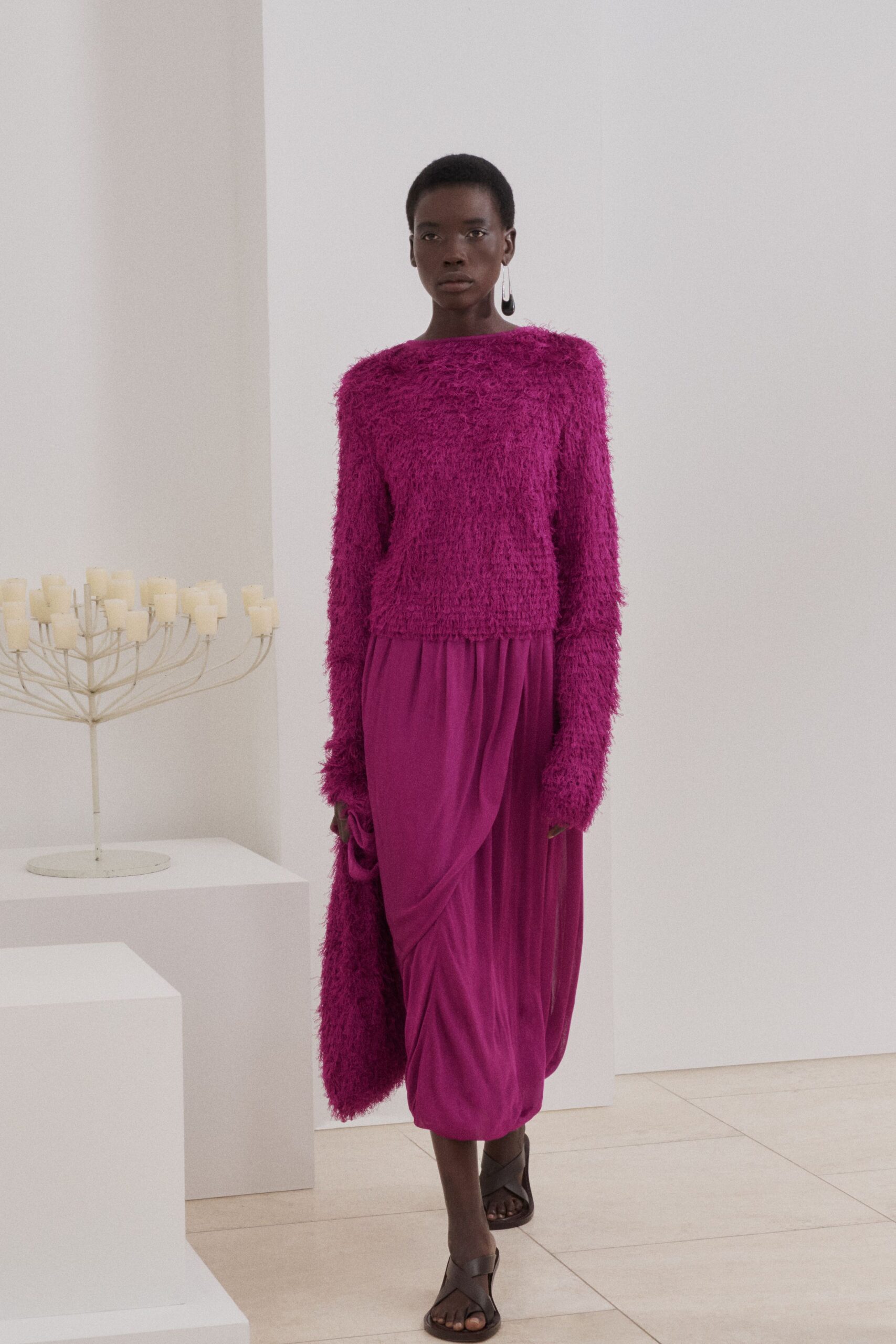 Fuchsia eyelash-knit sweater with a wide bateau neck and long shaggy sleeves. Matching fuzzy scarf bunched in one hand. Magenta jersey skirt softly draped and twisted across the hips. Dark leather crossover sandals. Walking through a candle showroom before anything has been lit.