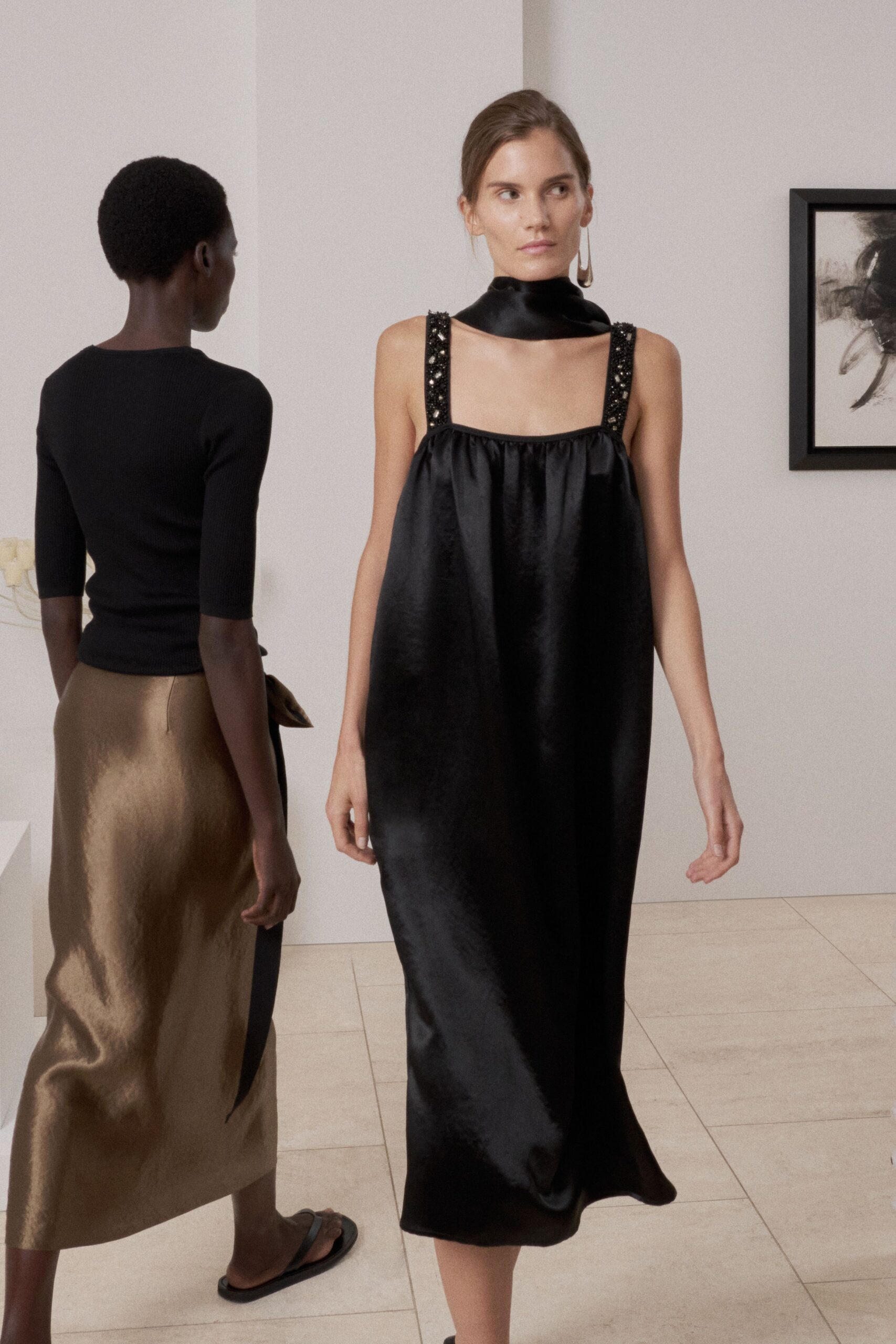 Black satin collar wrapped close, cut open across the collarbones. Inky satin column dress with beaded straps and a softly gathered square neckline. Long drop earring in dull gold. Crossing a quiet gallery during the collector's breakfast.