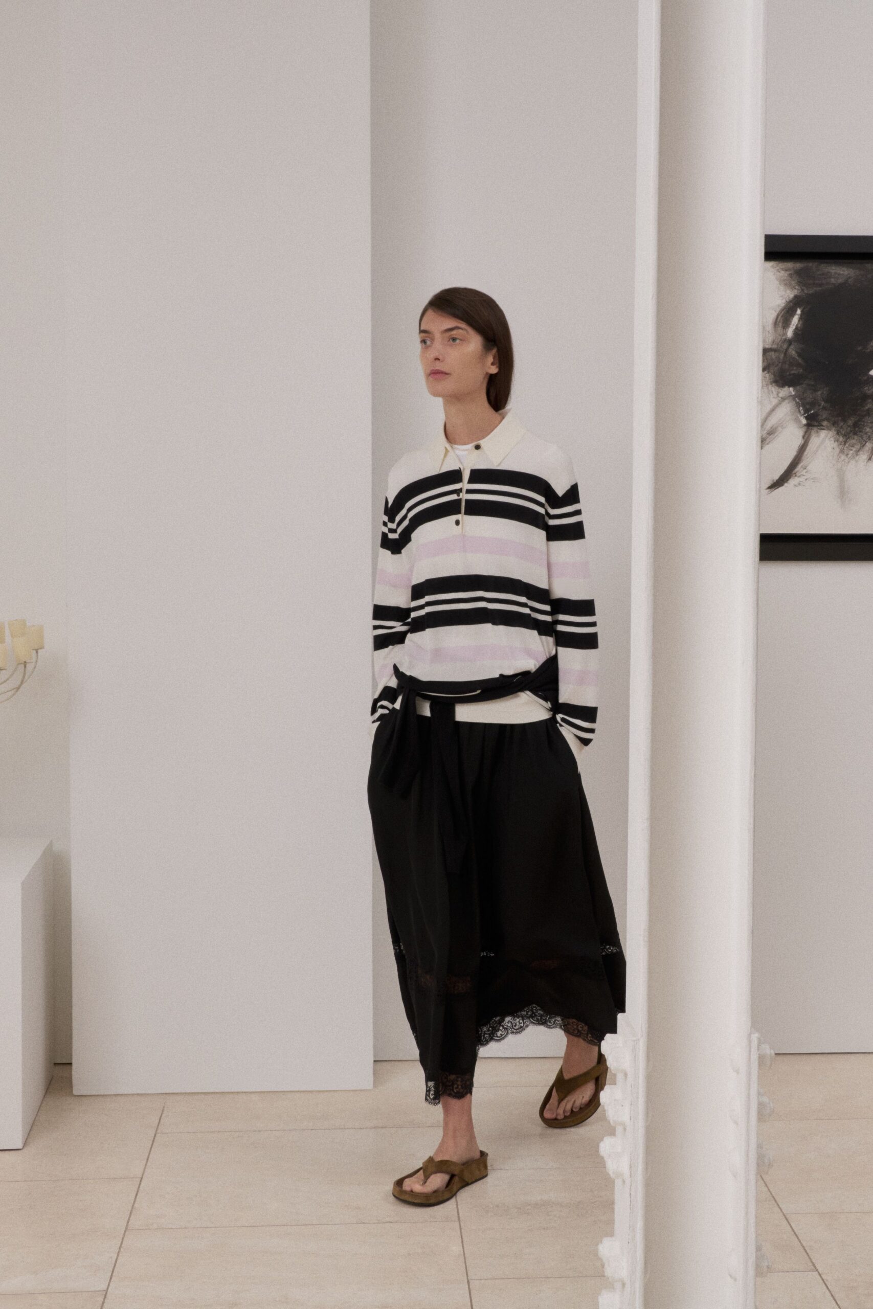 Fine white knit polo with black and blush bands, third button undone. Black gathered midi skirt with a tied overskirt panel and lace hem. Moss suede thong sandals on thick cork soles. Walking through a quiet house rental before anyone else wakes.