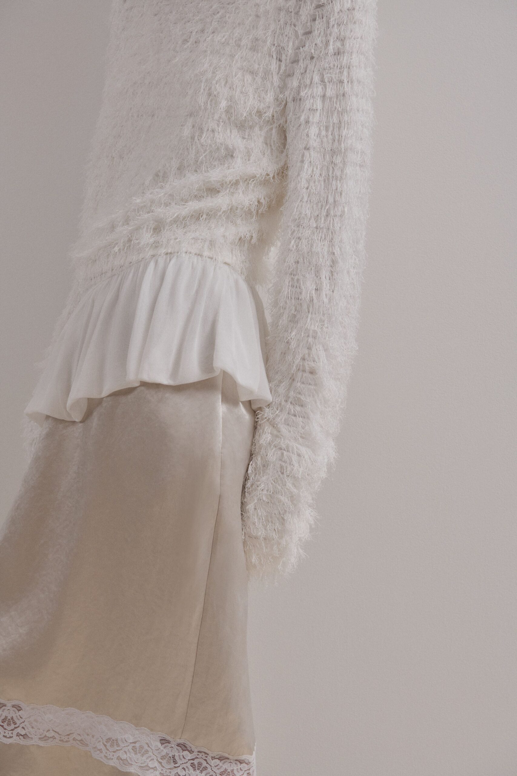 Ivory eyelash-knit sweater, loose ribs showing through the sleeve. Soft white peplum flounce breaking at the waist seam. Champagne satin slip skirt finished with scalloped lace at the hem. Sunday morning in a rental kitchen, coffee cooling beside the sink.