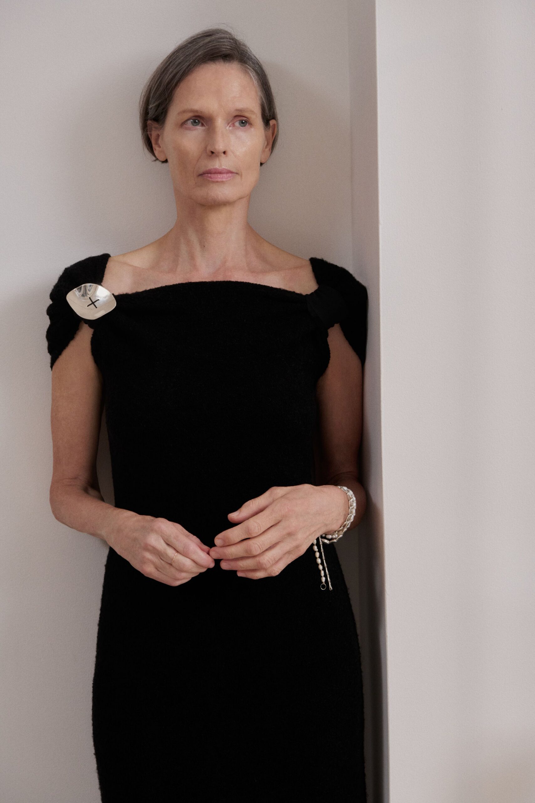 Black boucle column dress with a straight neckline and knotted shoulder straps. Crumpled silver disc brooch, marked with a hand-drawn black cross. Chain bracelet with a pearl-ended drop hanging past the wrist. Thin gold band on the left hand. Standing in a quiet hallway after everyone else has left dinner.