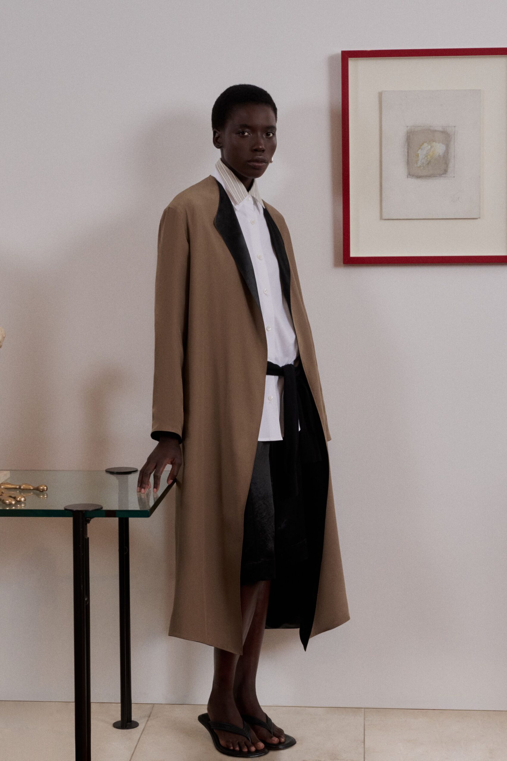 Long camel coat with black satin lapels and a loose self-tie. White button-front shirt with a narrow beige banker-stripe collar. Black below-knee skirt catching a faint moire sheen. Flat black thong sandals with ridged straps. For walking through a quiet house after everyone else has gone to bed.