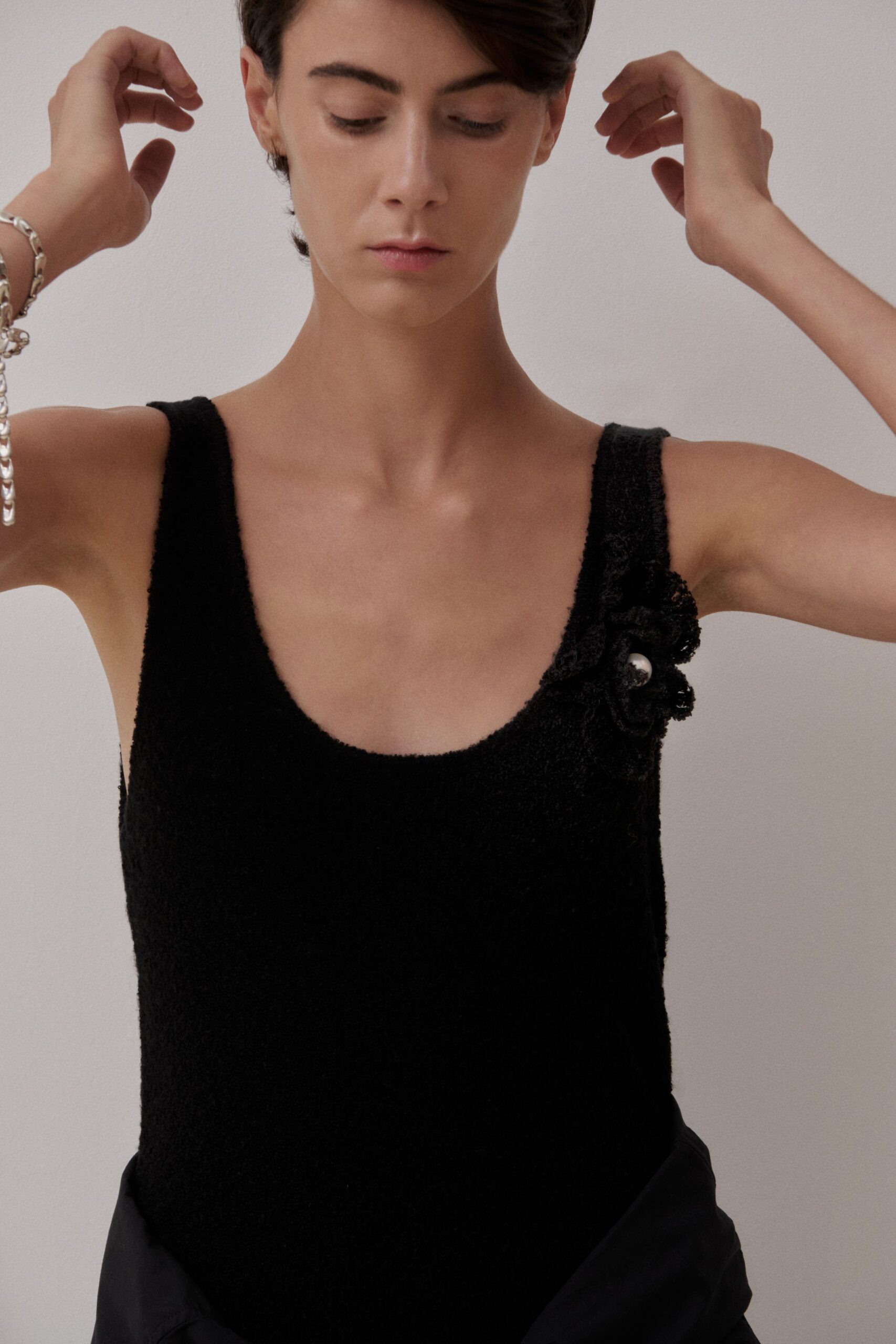 Heavy silver curb chain bracelet slipping toward the wrist bone. Black boucle-knit tank dress with a low scoop neck. Lace rosette at one strap, pinned with a single pearl bead. Morning of a civil ceremony, steam still on the bathroom mirror.