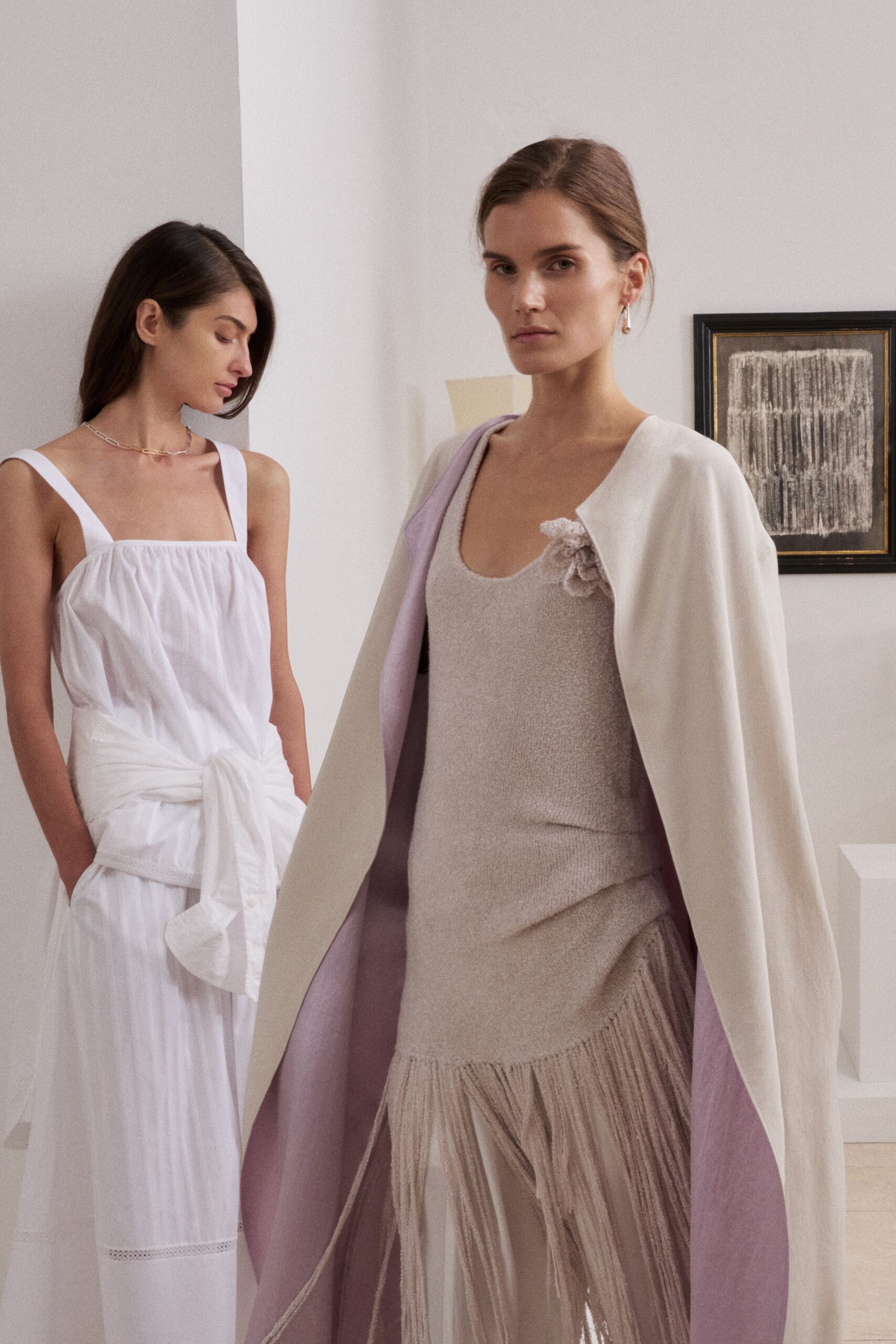 Ivory cape with a lilac underside, hanging open without closures. Pale heather knit dress with a raw-edged flower at one strap. Long tonal fringe spilling from an uneven dropped panel. Quiet hotel suite, dressed before the ceremony while the flowers are downstairs.