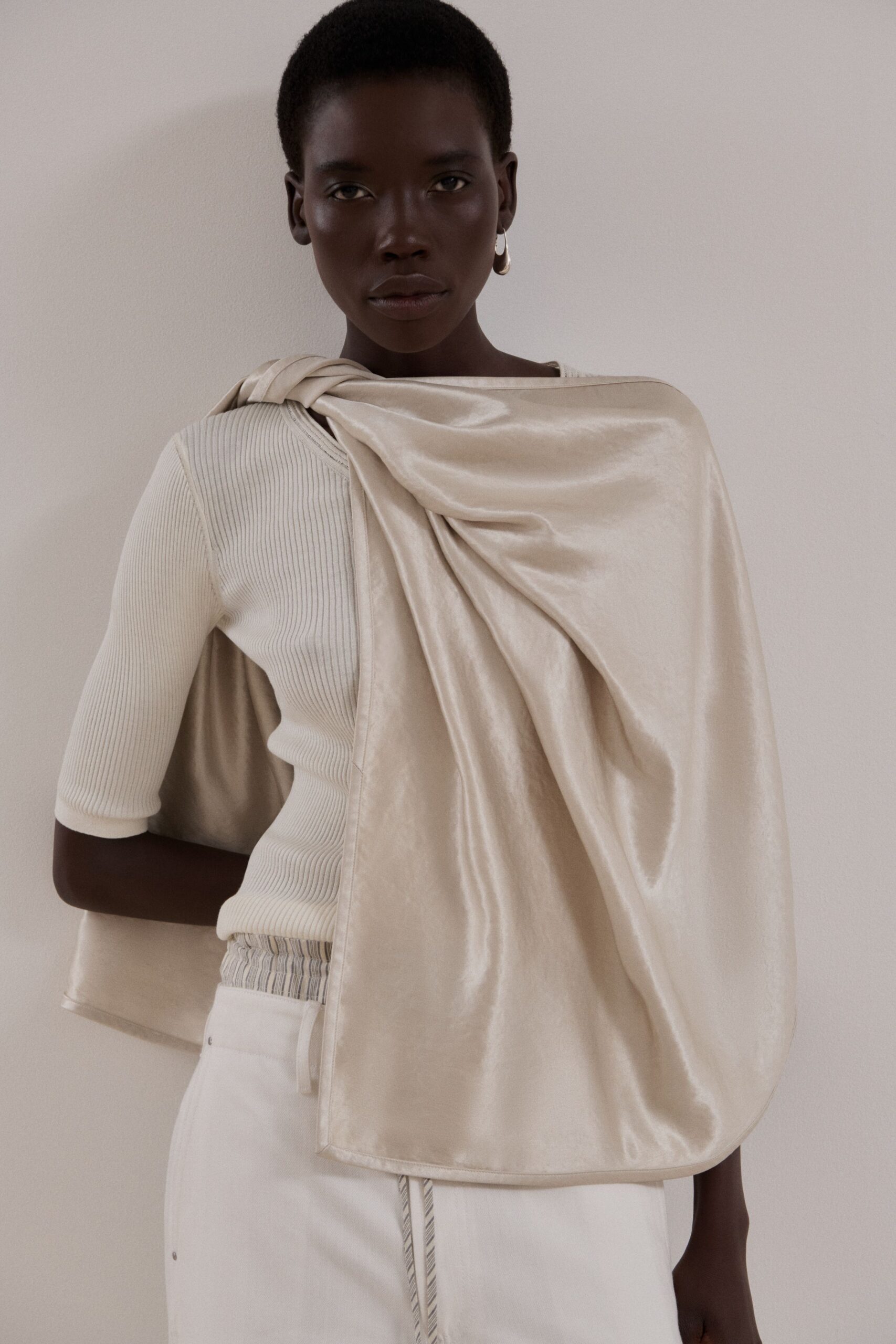 Single small silver hoop. Ivory ribbed knit top with elbow sleeves and a curved cutout side. Champagne satin panel knotted at one shoulder, falling blanket-like across the torso. White column skirt with striped drawcord hanging past the waist seam. Museum atrium at opening hour, coat still folded over one arm.