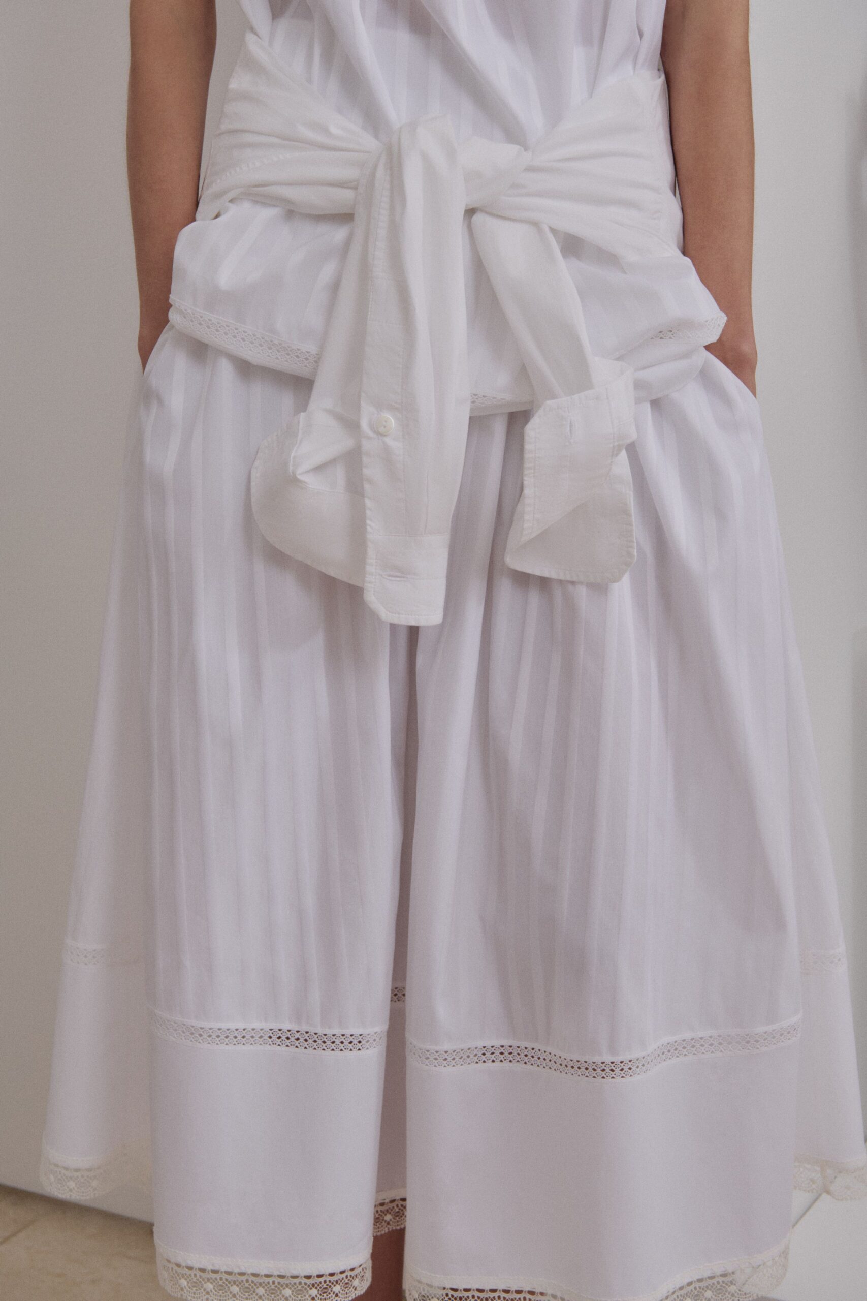 Sheer white voile blouse falling in long vertical folds. White cotton shirt knotted hard at the waist, one button showing. Wide white culottes banded with narrow lace insertion near the hem. For crossing a tiled courtyard just after the sheets came off the line.