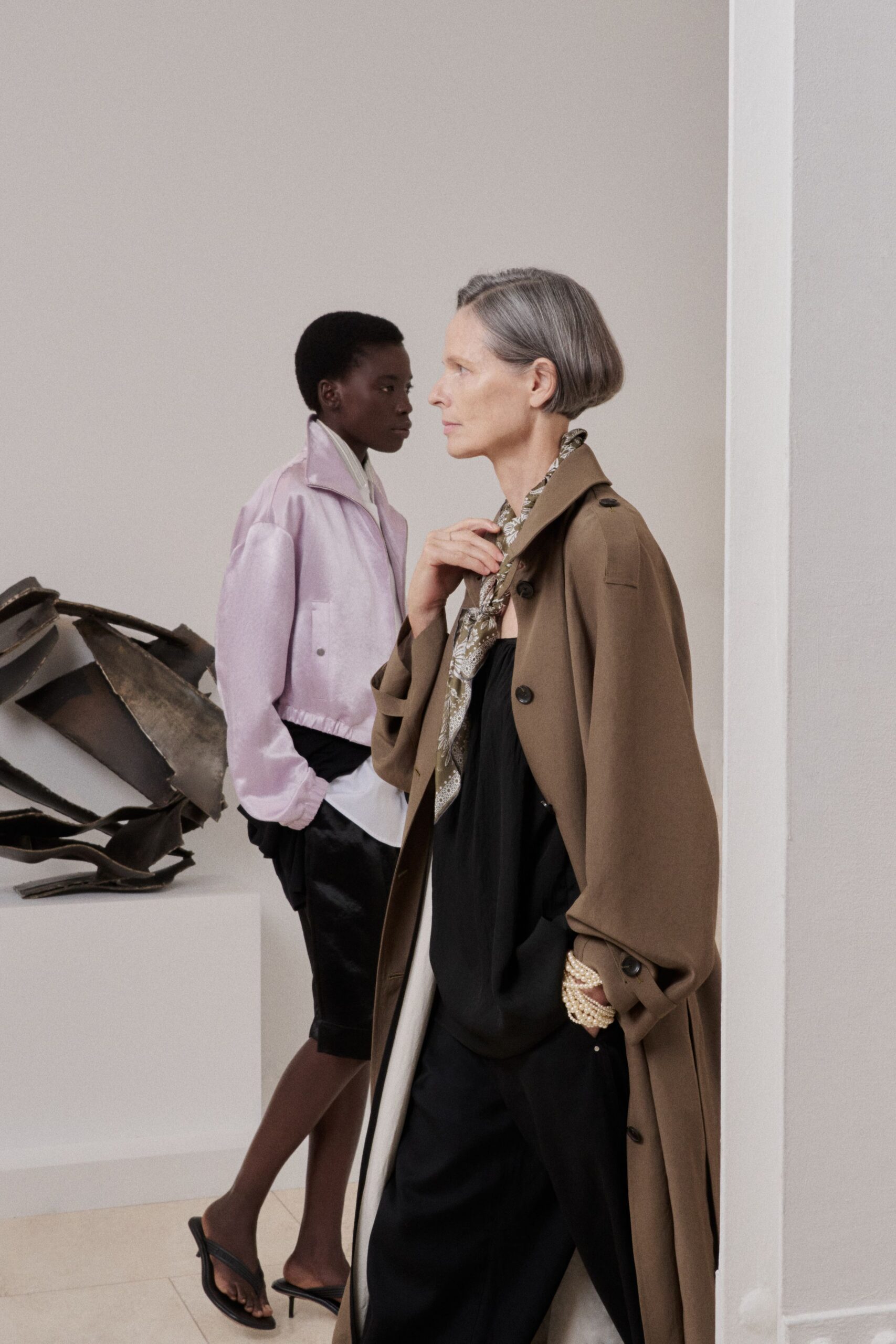 Taupe trench in dense twill, sleeves pushed over the knuckles. Olive paisley silk scarf knotted close at the throat. Black gathered camisole, soft folds pooling above the waist. Black wide-leg trousers with a fluid, light-catching drape. Clustered pearl strands at the wrist. Crossing a quiet museum atrium before the doors open.