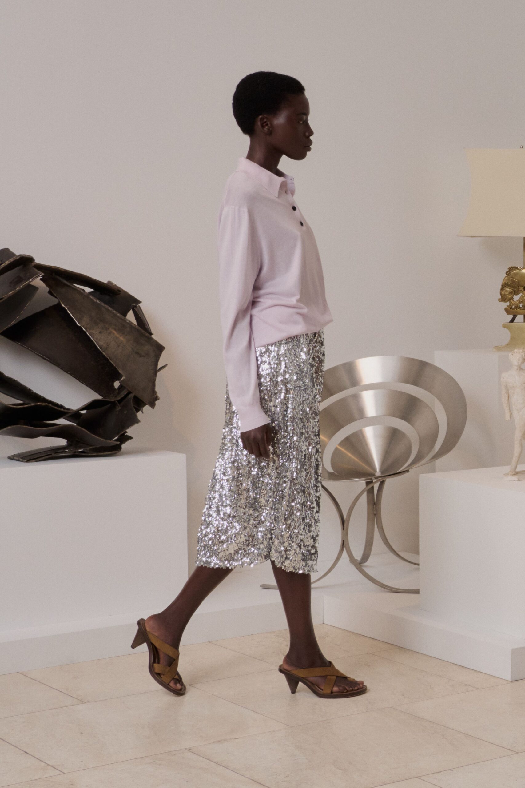 Pale blush knit polo with a soft drop shoulder and dark buttons. Silver sequin midi skirt, densely pailletted and swinging in a straight column. Olive leather cross-strap mules on a low flared heel. Crossing a sculpture garden terrace before the champagne is poured.