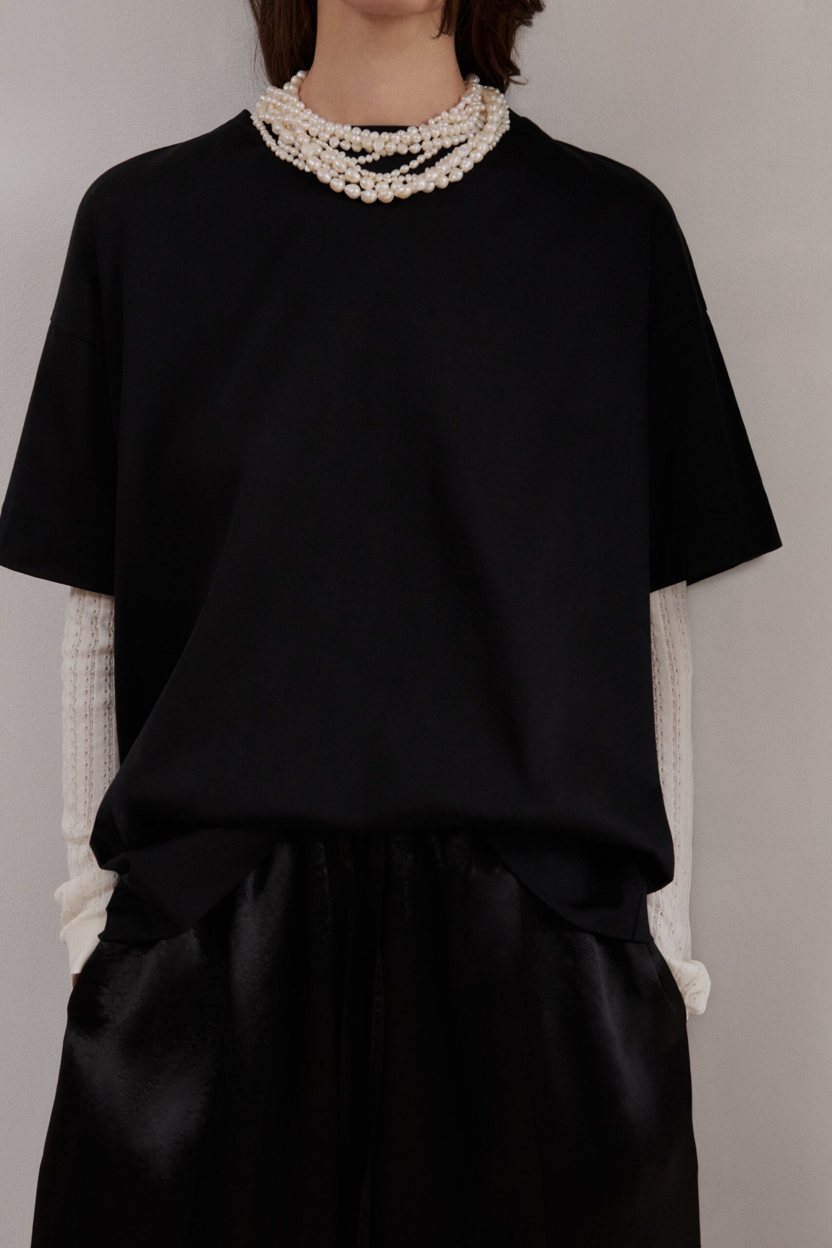 Four uneven strands of baroque pearls piled close at the throat. Black oversized tee in smooth jersey, sleeves cut wide at the elbow. Cream pointelle knit sleeves peeking from underneath. Black satin trousers with a soft, light-catching sheen. For picking up flowers in the morning before changing for the memorial.