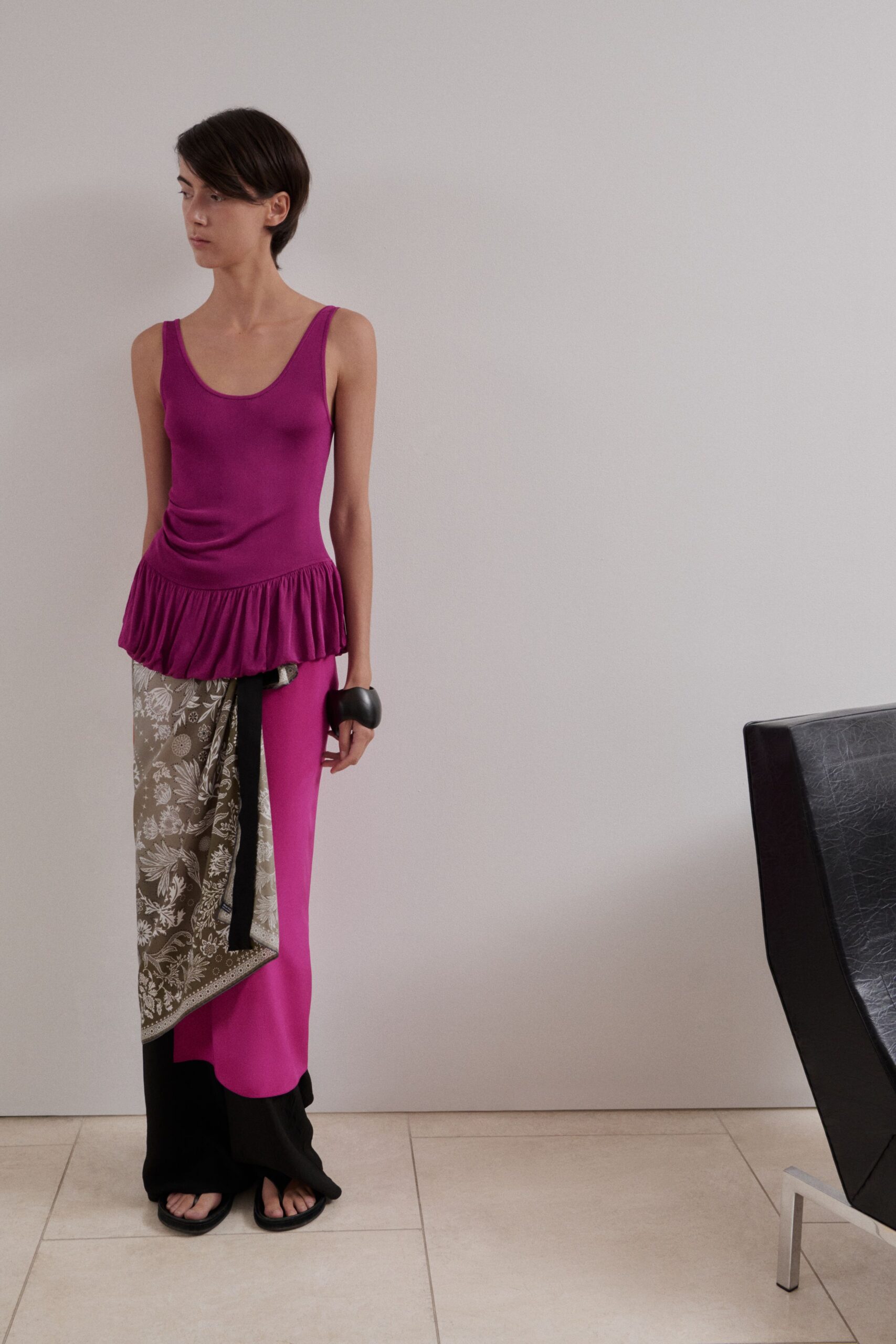 Magenta jersey tank dress with a dropped waist and soft peplum flounce. Taupe floral scarf panel tied at the hip, one black tail hanging long. Black trousers pooling over the foot in fluid crepe. Oversized black resin cuff. Flat black thong sandals. Walking across cool tile before lunch, house still quiet after guests left.