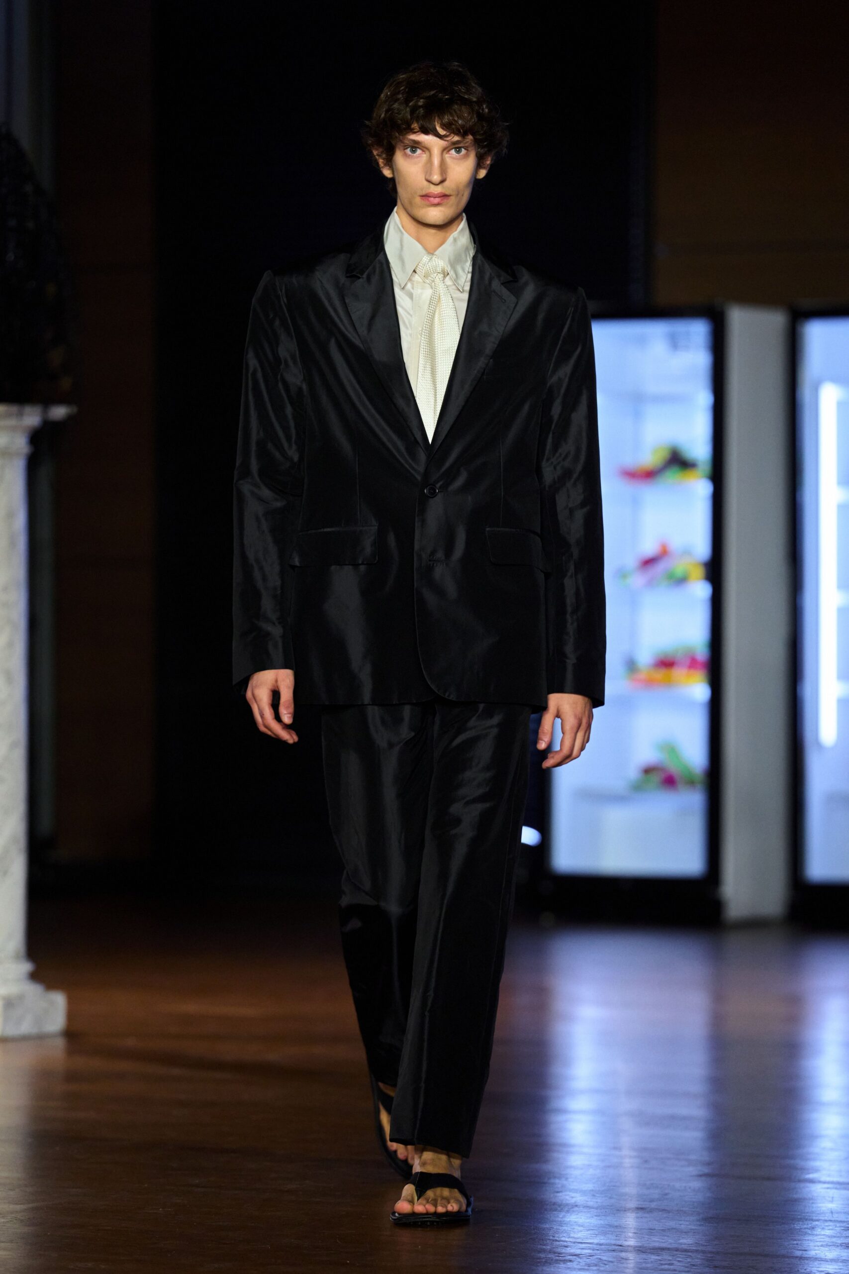 Black satin blazer with narrow lapels and a single low button. Pale grey shirt, collar points stretched long under the jacket. Ivory tie in tiny textured weave. Matching satin trousers breaking clean over the foot. Black flip-flop sandals. Walking through a hotel lobby after midnight, still dressed for the wedding.