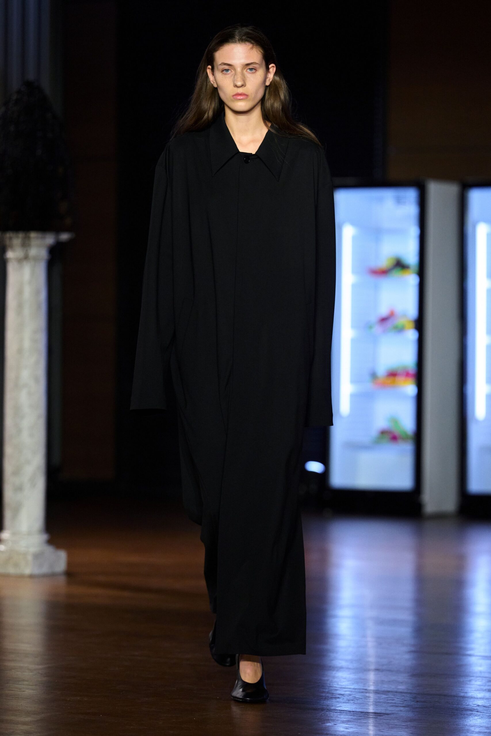 Black floor-length coatdress in fluid crepe, placket hidden under a pointed collar. Black leather pumps with a blunt square toe. Crossing a silent museum foyer after the cleaners have finished.