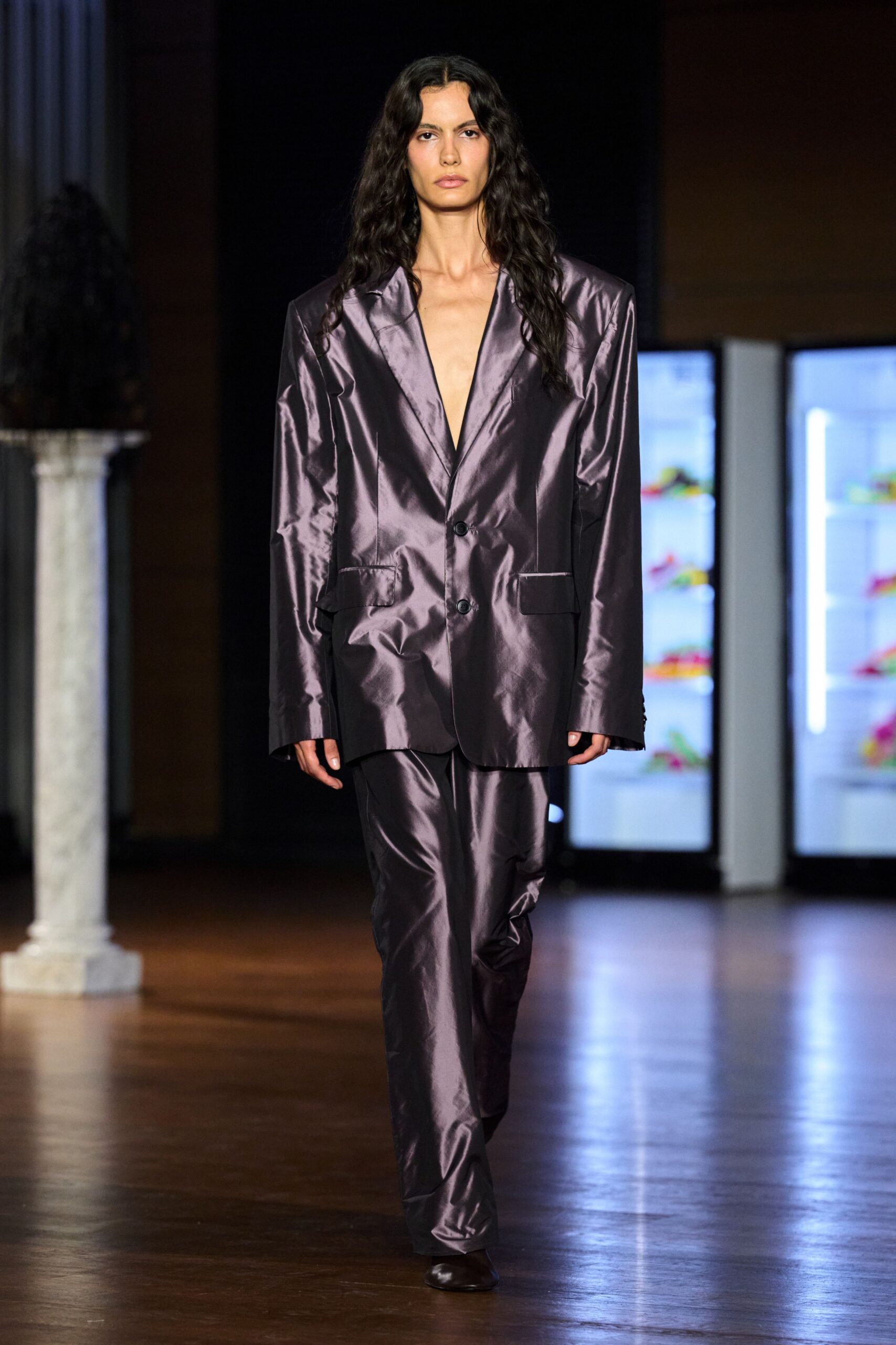 Oversized violet-grey satin blazer, two black buttons and a plunging bare chest. Matching satin trousers, softly crushed through the shin. Dark square-toe leather shoes. Walking past the hotel lobby fridges after midnight for ice.