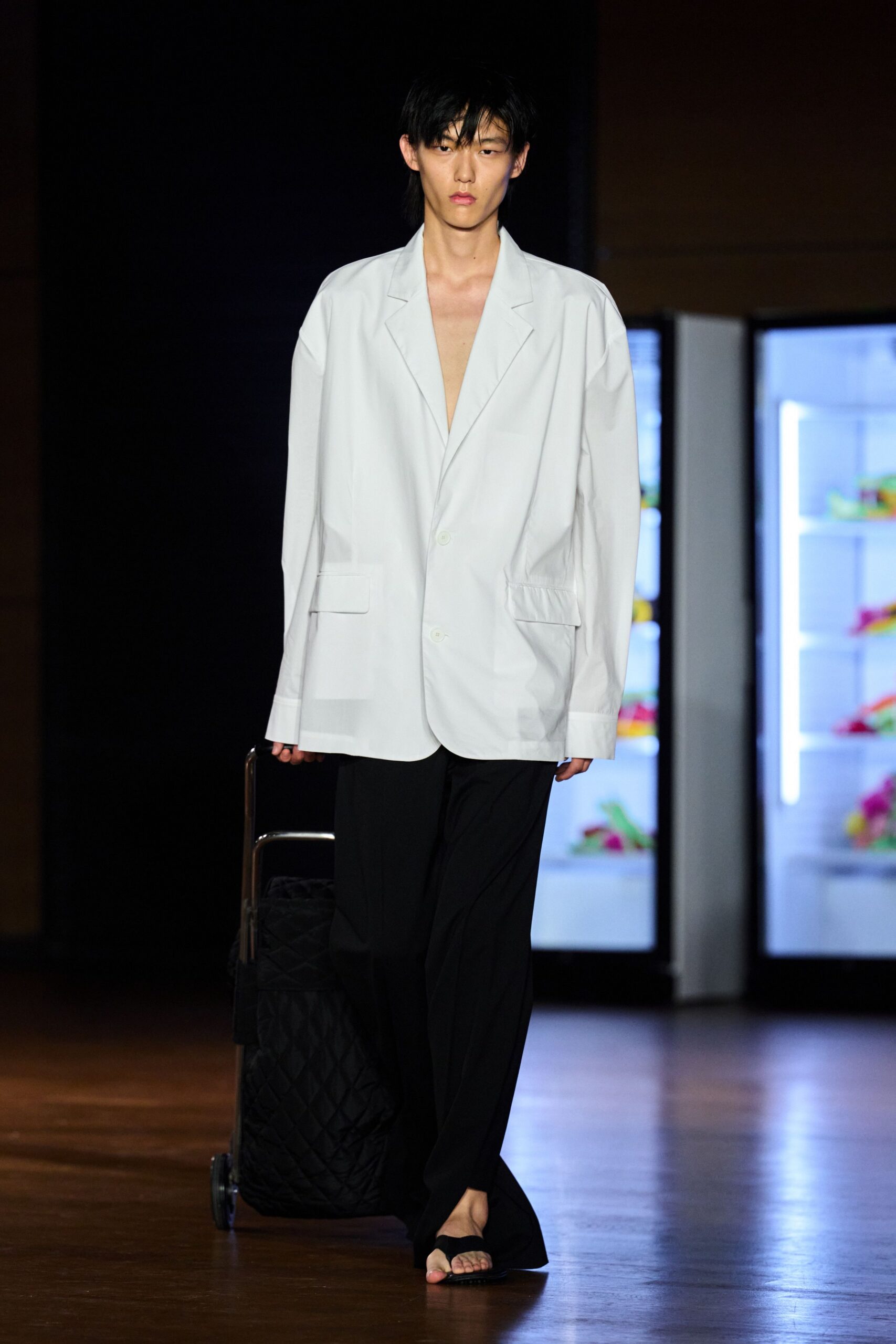 Pale blue cotton blazer cut oversized, deep V left bare to the waist. Black fluid trousers breaking softly over the instep. Flat black slides with a wide suede strap. Black quilted suitcase dragged beside the leg. For the first hotel corridor after a red-eye, minibar light still humming.