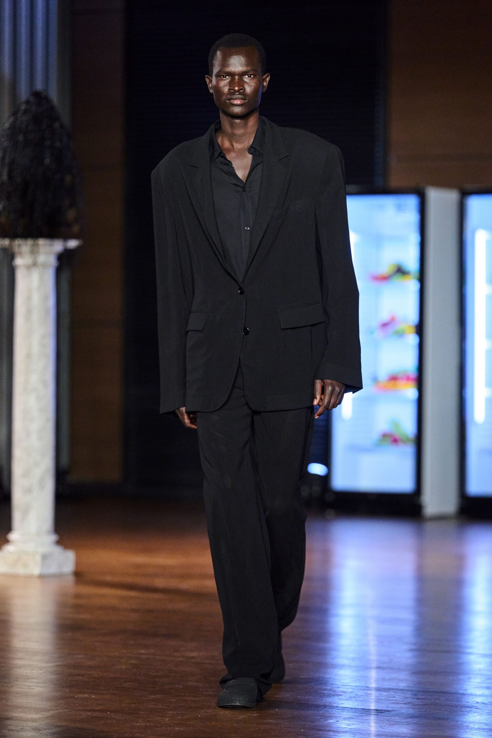 Black suiting blazer cut loose through the shoulder, two-button front. Black shirt in fluid matte cloth, top buttons left casually open. Straight black trousers with a soft break pooling over the shoe. Square-toe black shoes. For crossing a hotel lobby before sunrise, keycard already in hand.