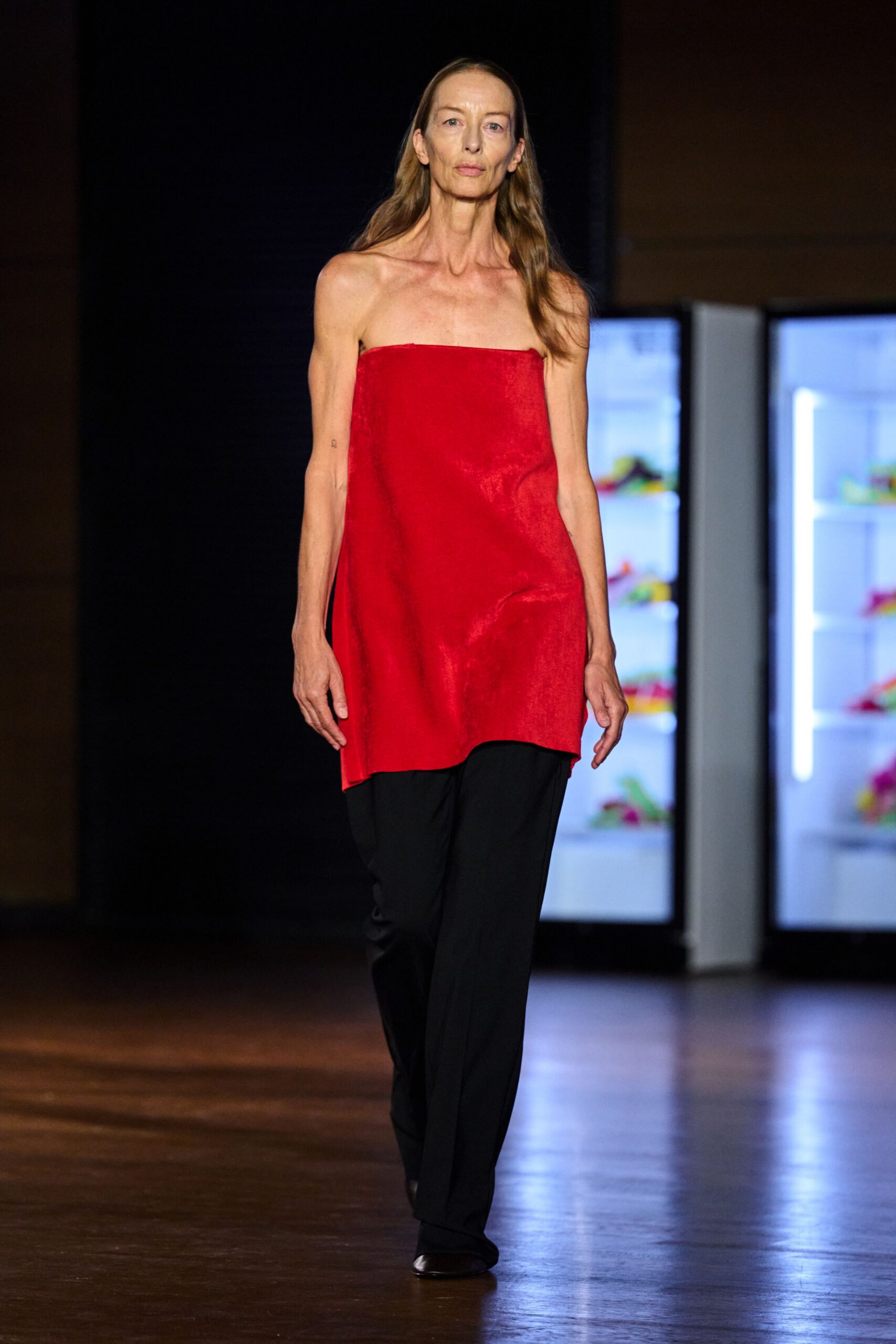 Scarlet strapless tunic in lustrous satin, hem dipping lower at one side. Black fluid trousers breaking softly over the shoe. Dark square-toe flats with a low shine. For the quiet table in a hotel bar after midnight check-in.
