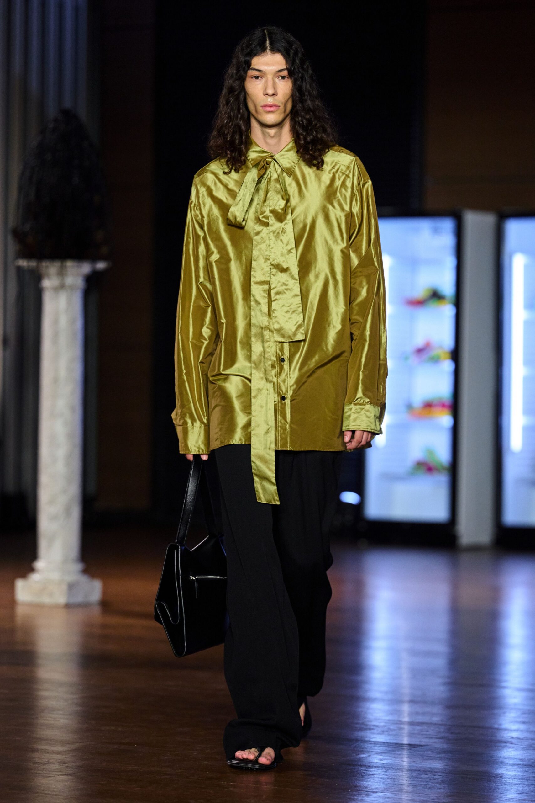 Olive satin blouse cut oversized, with a long scarf tie hanging past the placket. Black wide-leg trousers breaking softly over the foot. Structured black leather tote with a folded gusset. Black thong sandals, square at the toe. Walking through a museum after closing, still carrying the day's paperwork.
