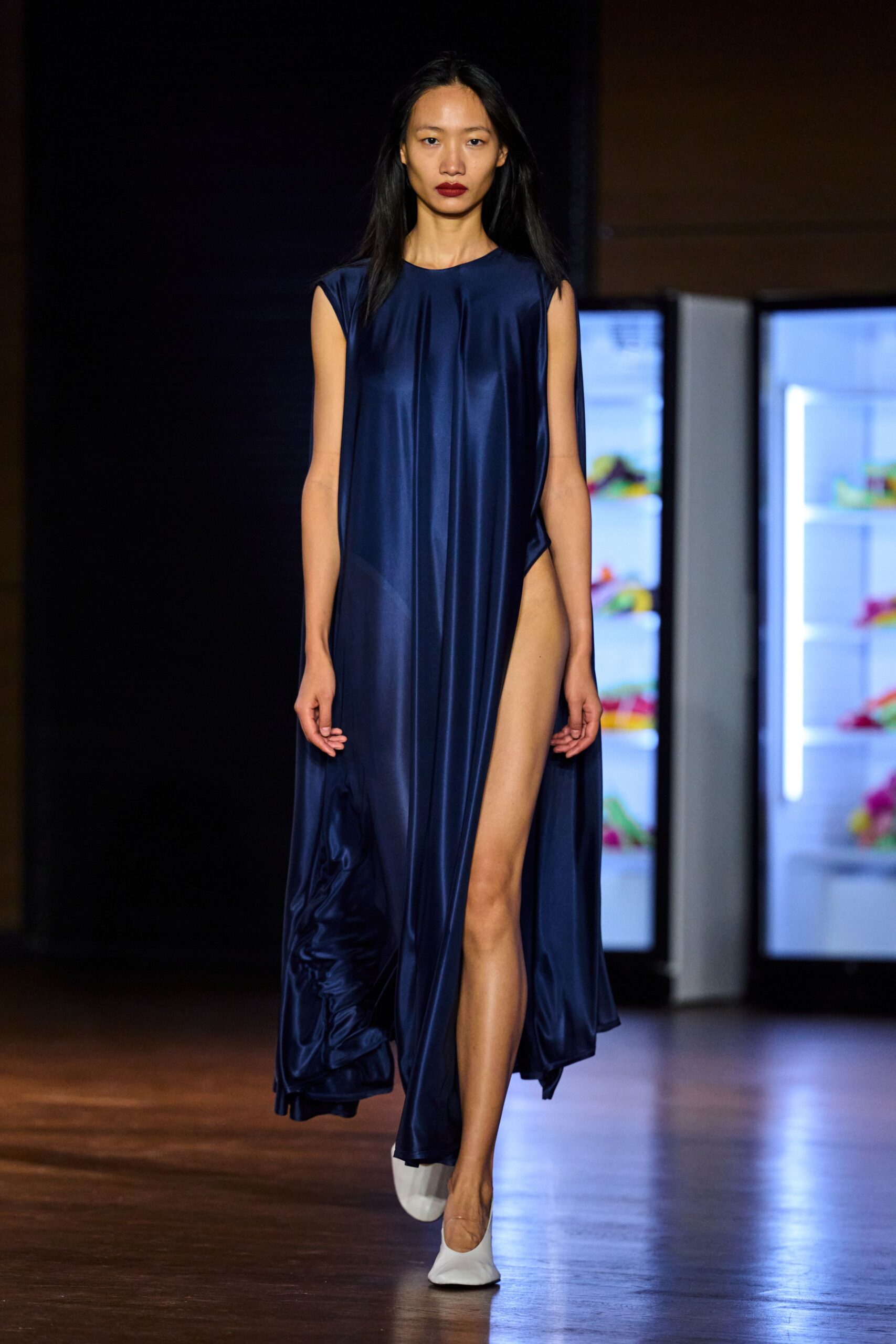 Ink-blue satin column dress, one side cut to the hip in a hanging panel. White square-toe flats. Crossing a hotel lobby at dawn before anyone has reset the lights.