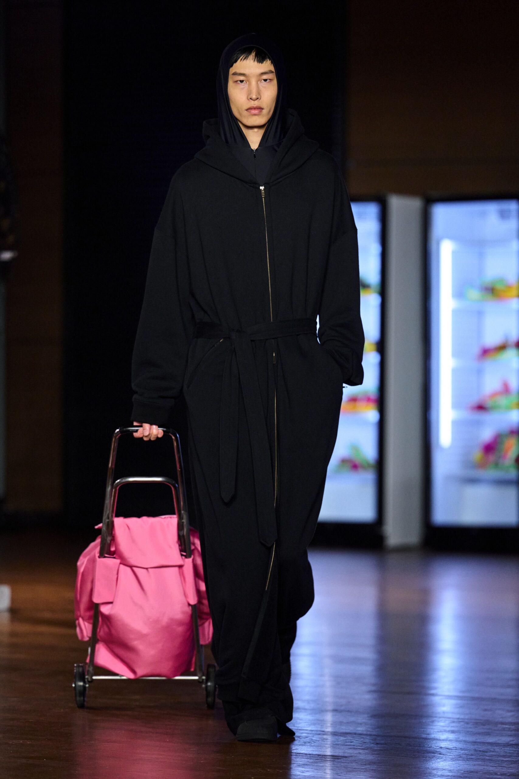 Black hood pulled close, zip hidden beneath a wrapped shawl collar. Long black jersey coat cinched with a self-tie belt. Black trousers pooling softly over the foot. Square-toed black shoes. Metal folding trolley hauling a bright pink nylon bag. For the cold walk back from the off-licence with too much to carry.