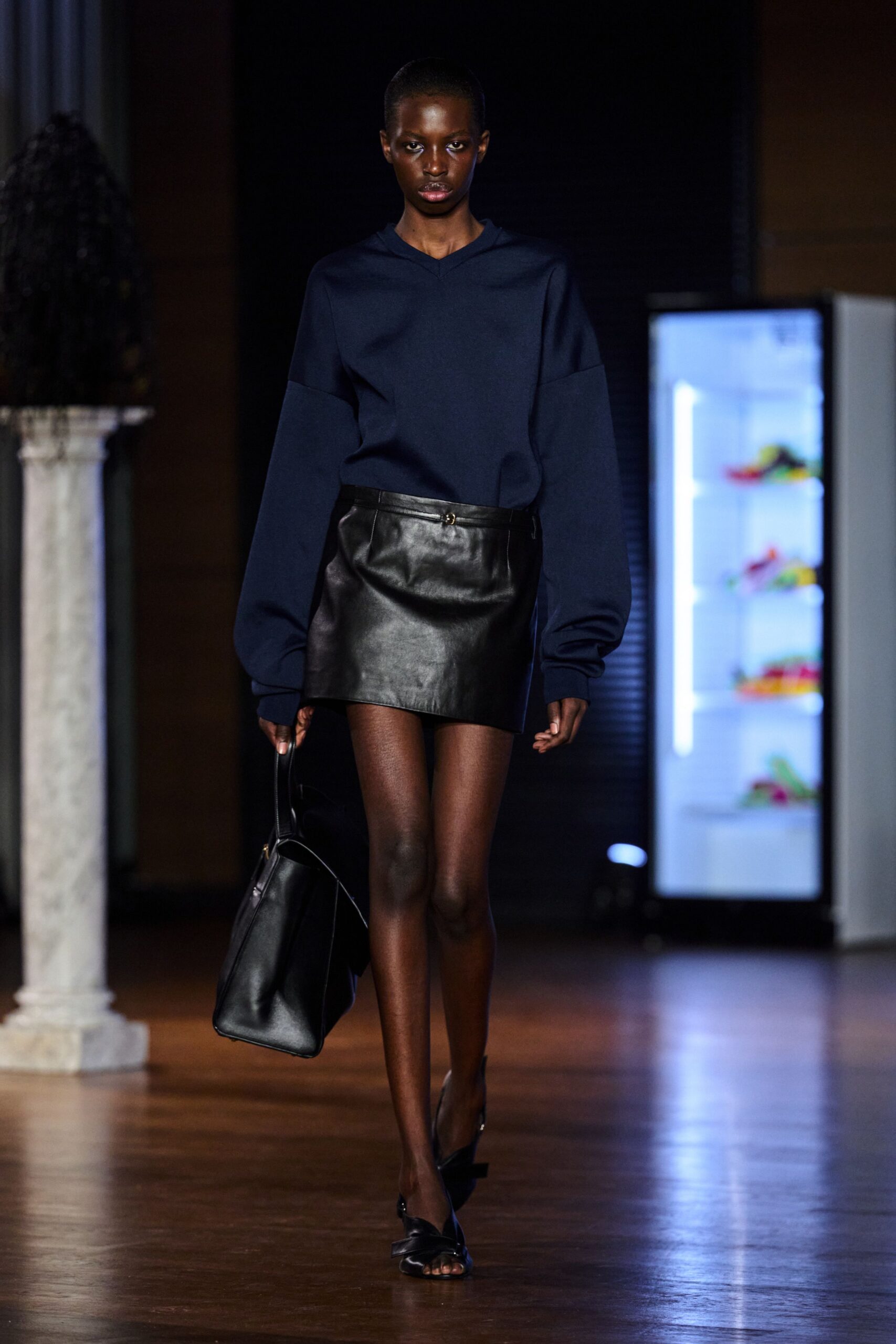 Navy knit top with a shallow V notch and dropped sleeves. Black leather mini skirt, wrapped slightly off-center with a tiny ring tab. Large black leather tote, boxy and soft at the base. Black open-toe heels with folded bow fronts. For crossing a museum atrium before the panel starts.