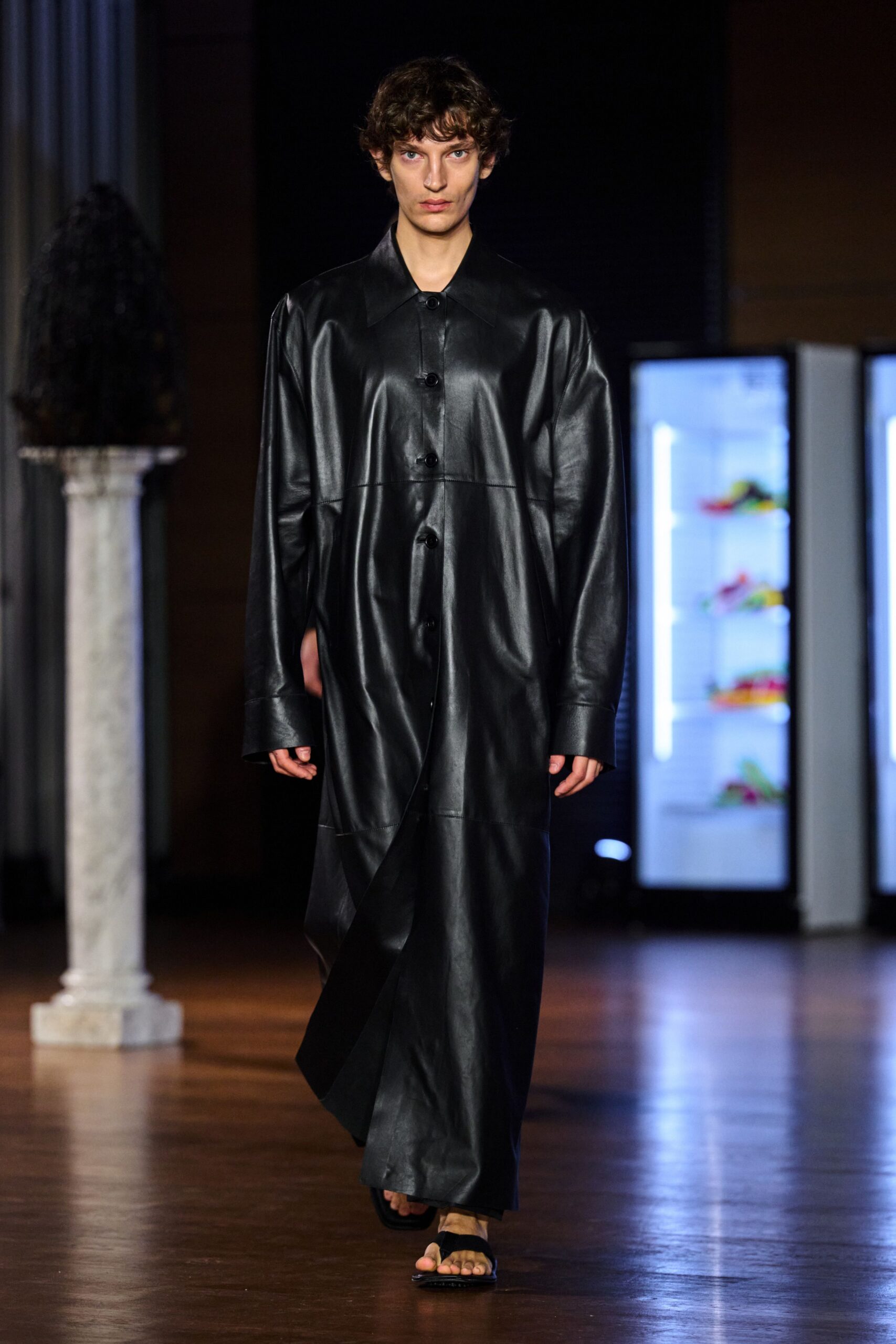 Black leather coatdress cut floor-length, with oversized collar and low glossy buttons. Bare thong sandals under the dragging hem. Crossing a museum hall after hours, floor wax still sharp in the air.