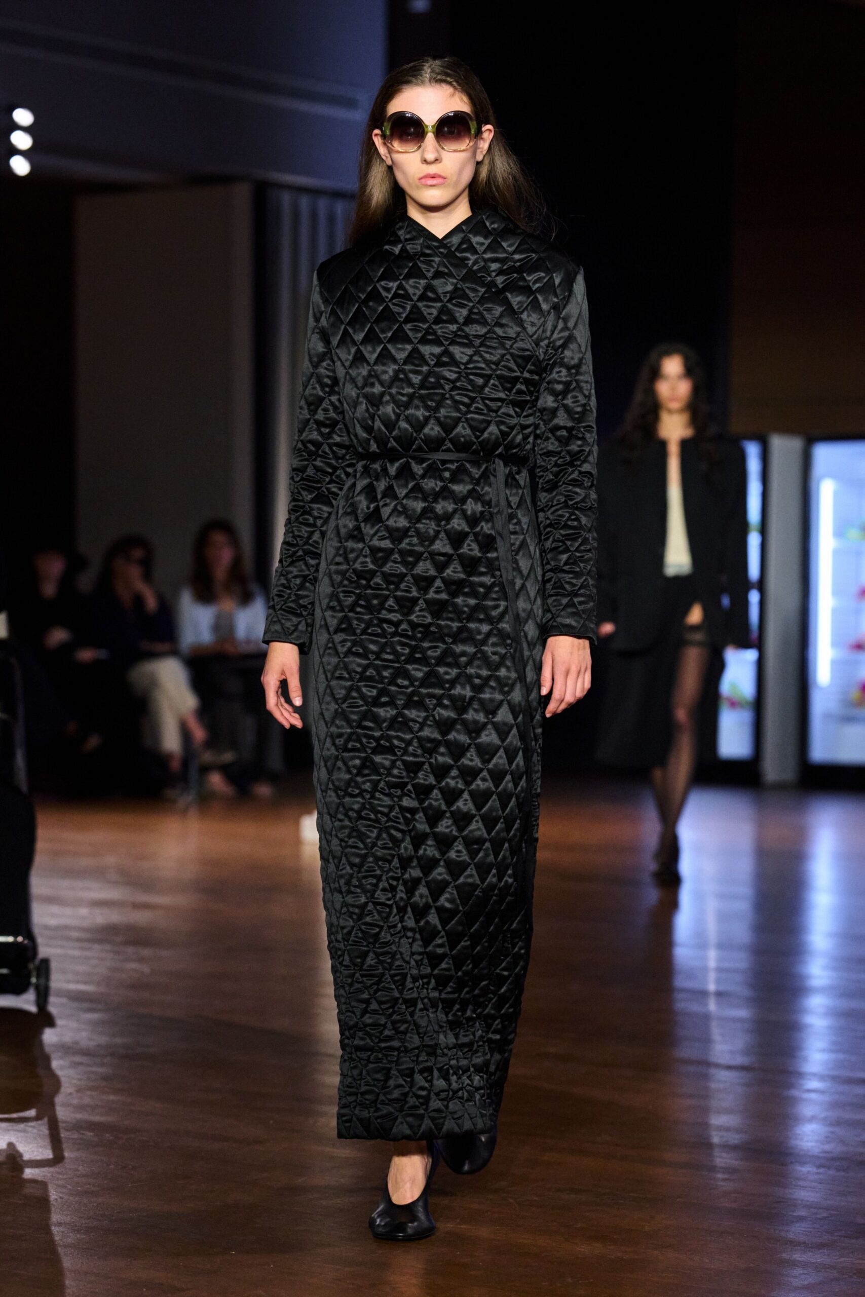 Oversized gradient sunglasses with thick olive-green arms. Black satin coat dress, diamond quilted and tied with a narrow self belt. Square-toe black leather flats. Crossing an echoing civic hall before the radiators kick on.