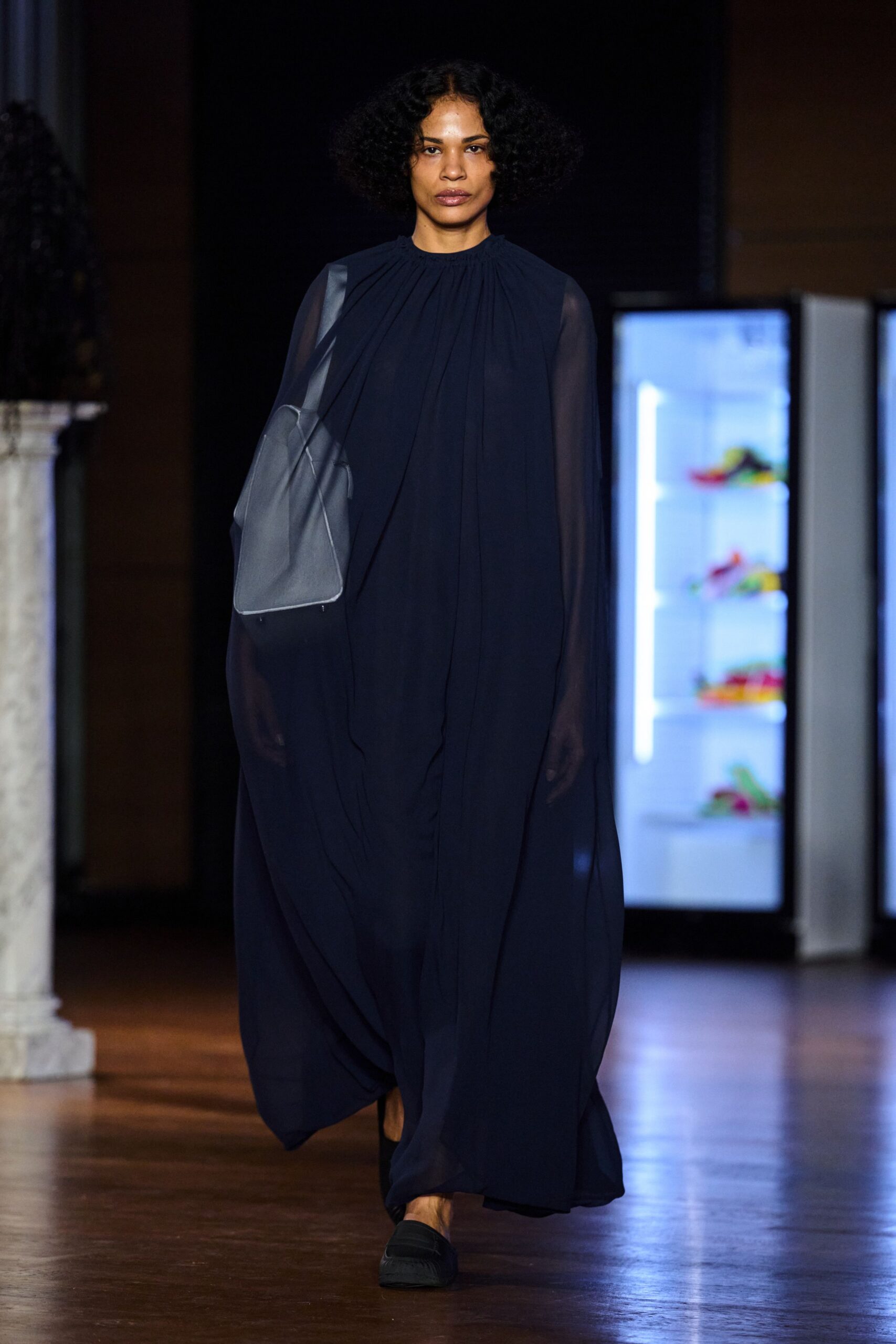 Ink-dark chiffon column dress, gathered tight at the neck and pooling low. Slate leather tote with a piped edge and flat shoulder strap. Black flats, square at the toe. Crossing a museum atrium before the rain lets up outside.
