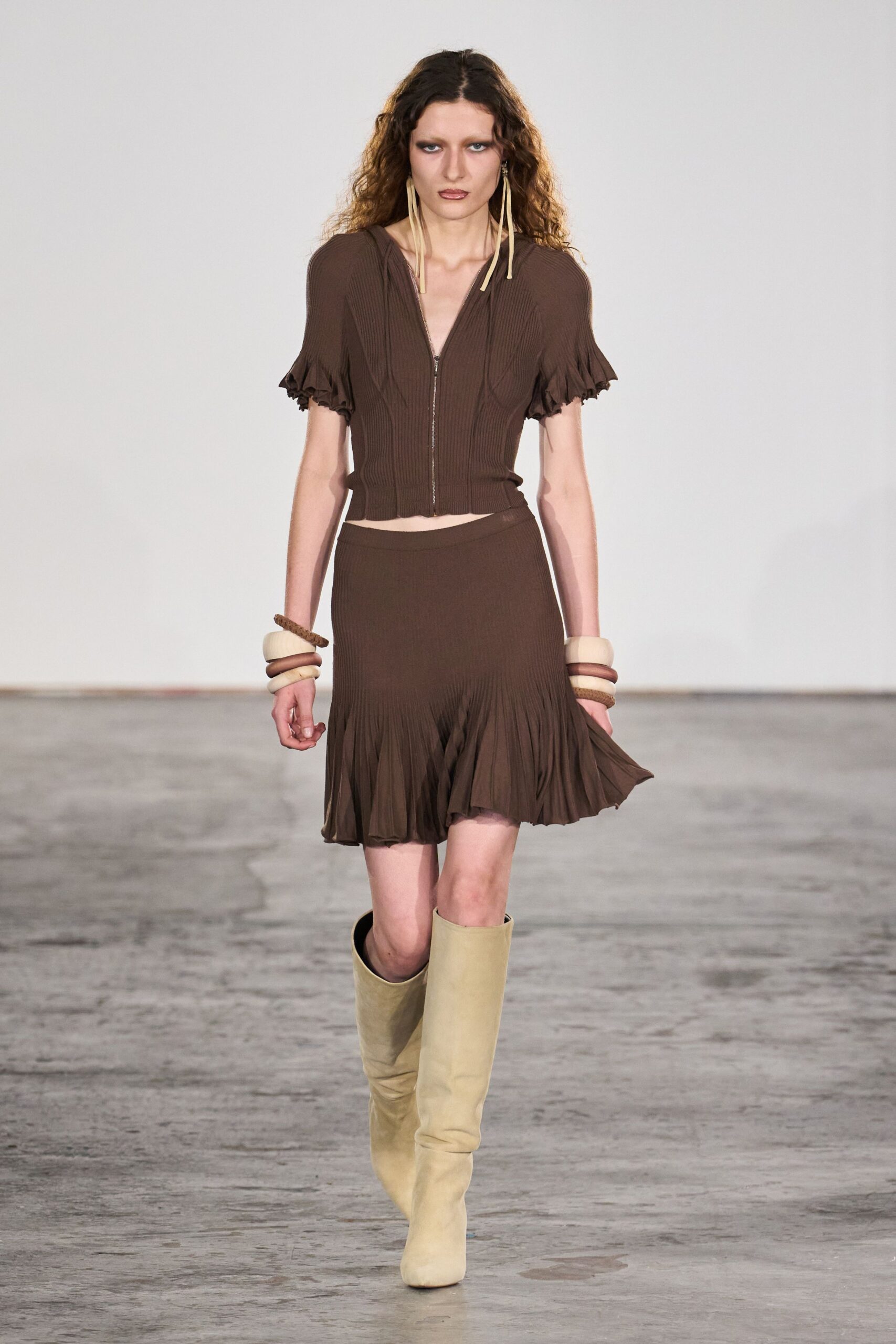 Long cream tassel earrings brushing the collarbone. Chocolate ribbed knit zip top with ruffled cap sleeves. Stacked ivory and brown bangles on both wrists. Matching ribbed mini skirt, hem released into soft pleats. Butter suede knee boots with blunt squared toes. Walking back from a hot spring before sunset dries your hair.