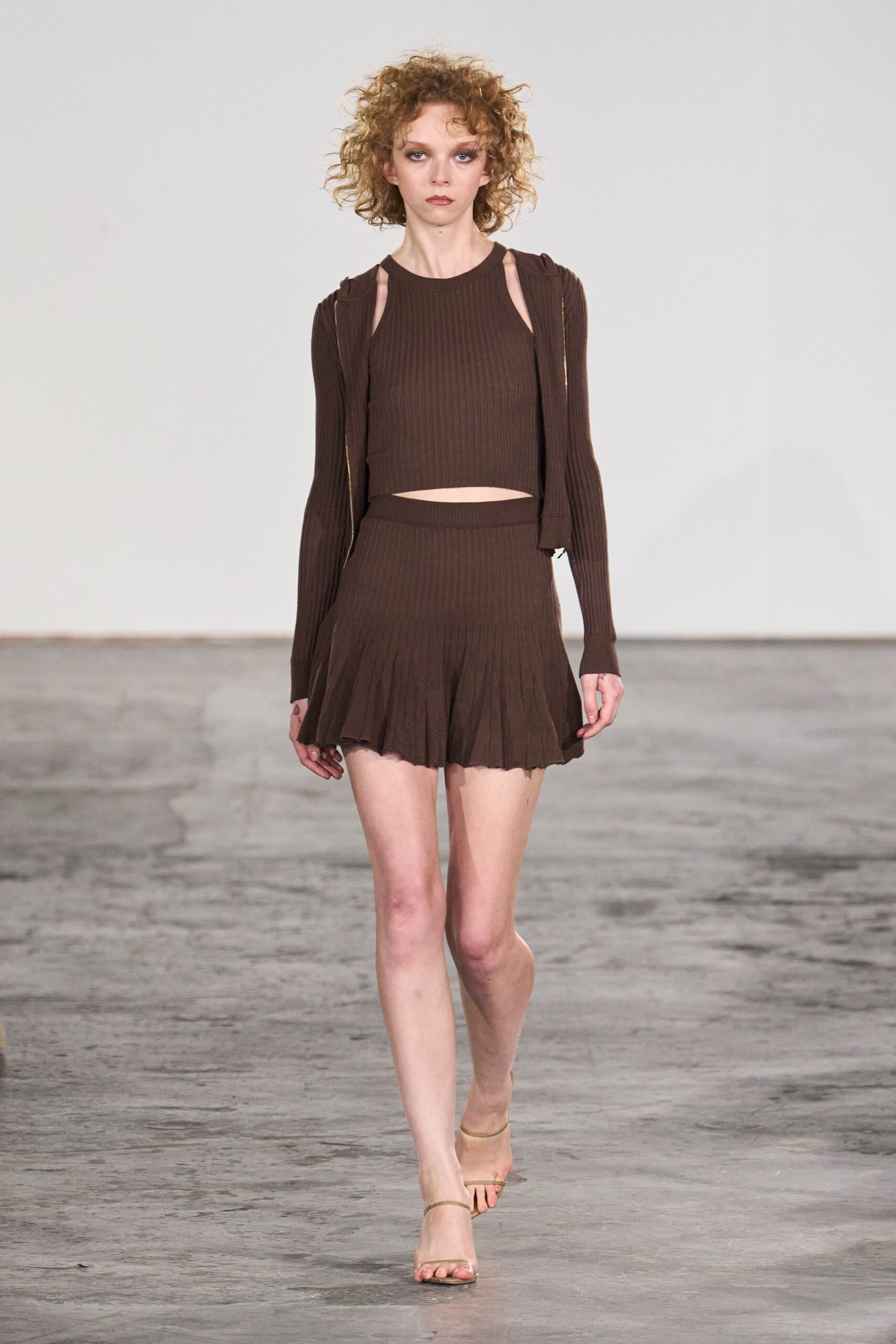 Chocolate rib-knit cardigan hanging open, long sleeves covering the palms. Matching ribbed crop top with narrow shoulder cutouts. Pleated knit mini flaring from a wide waistband, hem softly wavering. Barely there nude strap sandals. The first warm evening when nobody brought a jacket.