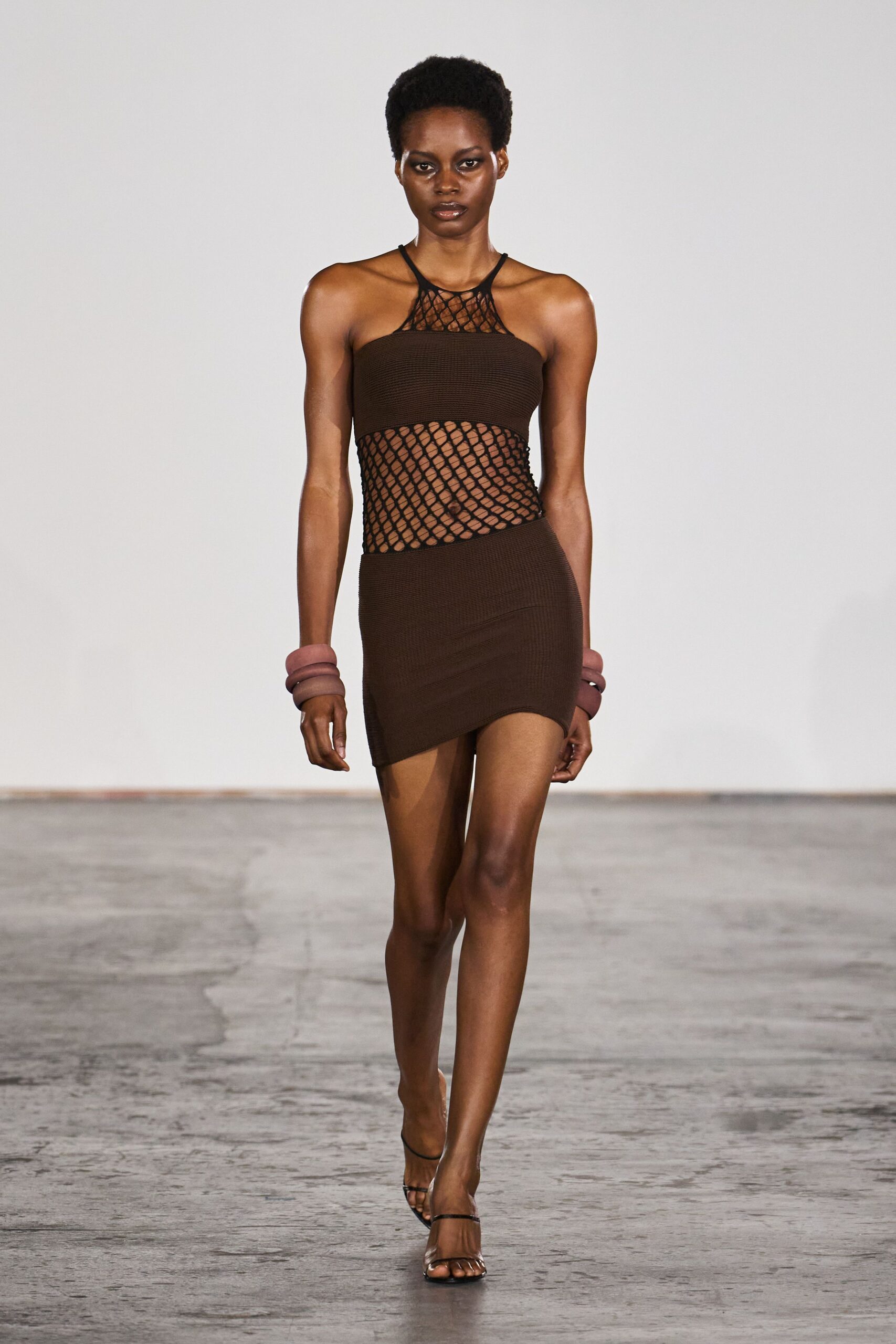 Espresso rib-knit mini dress, fishnet panels slicing through the bodice and waist. Stacked mauve cuffs at both wrists. Barely there black thong heels. Walking home from a beach bar after the heat still hasn't broken.