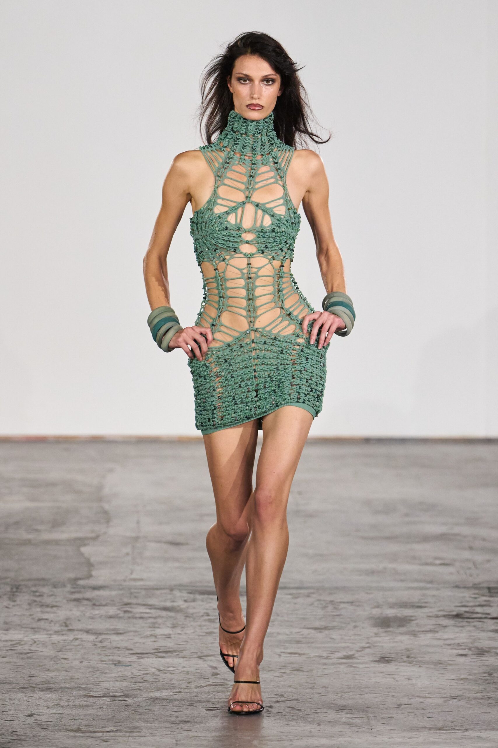 Seafoam openwork mini dress, laddered cords and knotted loops rising into a tall collar. Stacked translucent resin bangles, cloudy green at both wrists. Black barely-there sandals with a fine toe strap. Heat still radiating off the boardwalk after the souvenir shops shut.