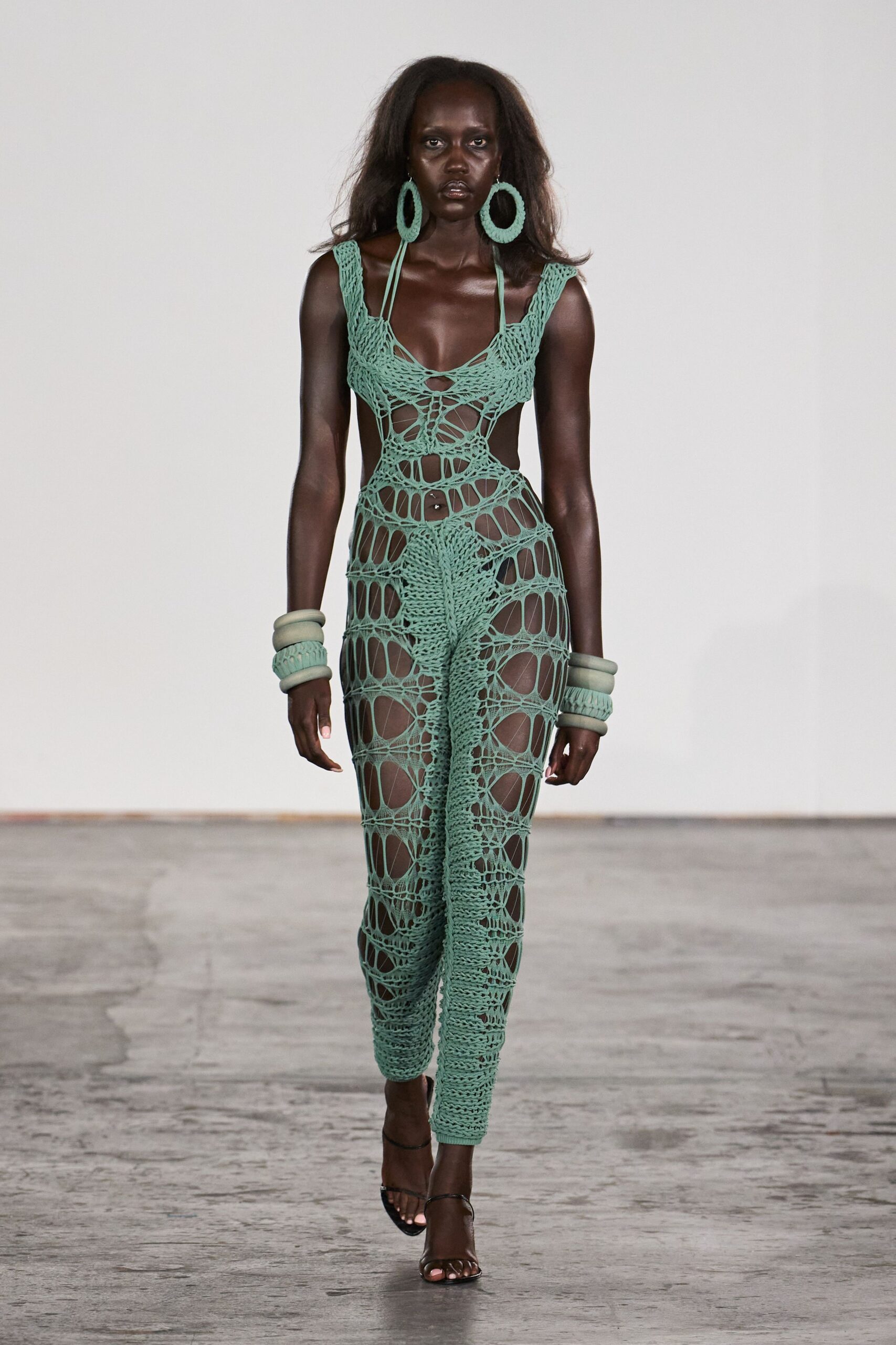 Oversized seafoam crochet hoops brushing the jaw. Seafoam openwork crochet jumpsuit, laddered wide at the hips and thighs. Stacked padded bangles in matching mint foam. Black barely-there sandals, one slim strap across the toes. Walking back from the beach with salt still drying on your shoulders.
