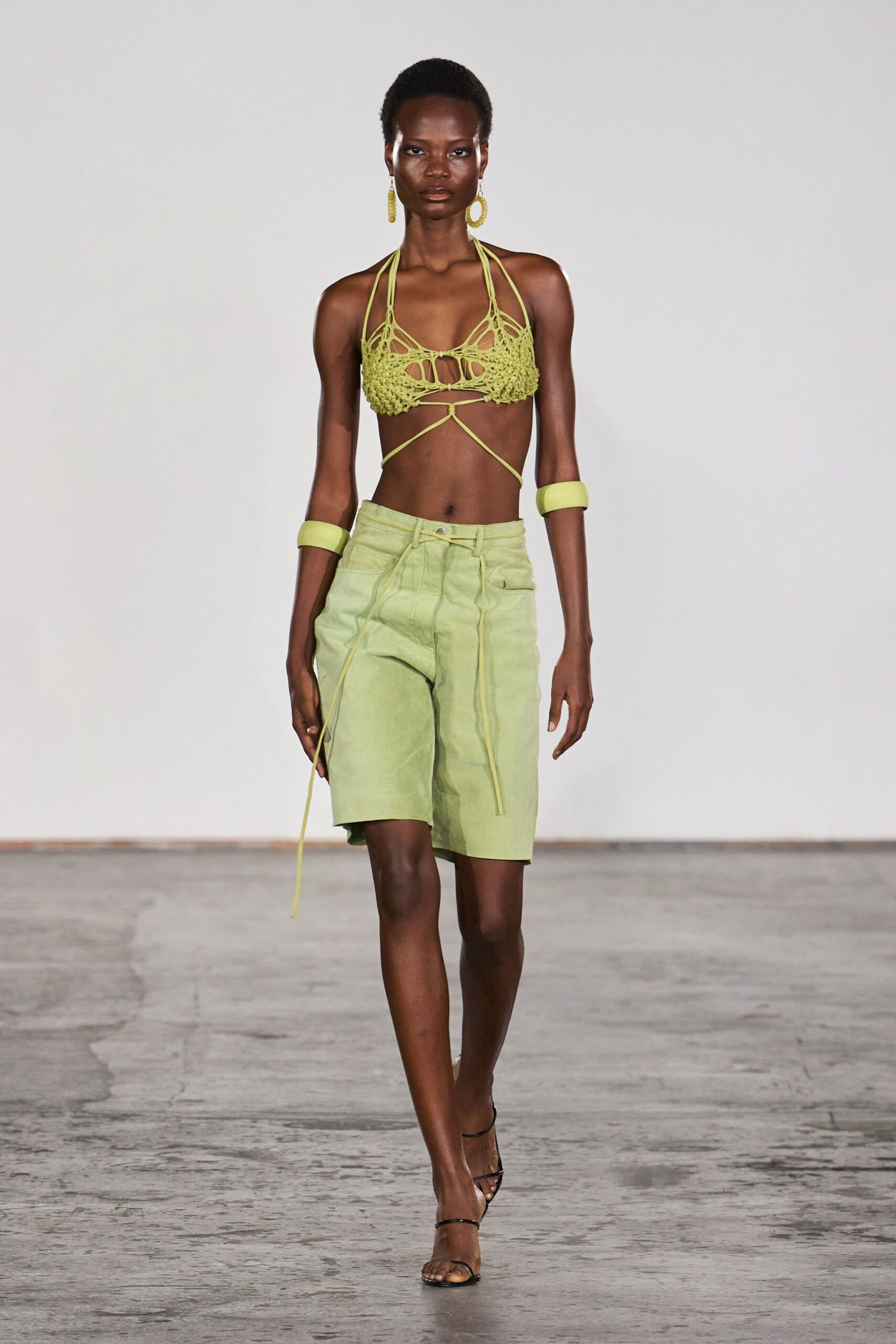 Celery crochet bralette, openwork cups tied with cords crossing the bare torso. Beaded chartreuse hoop drops. Foamlike lime bands at both forearms. Washed celery knee-length shorts, long drawstrings hanging past the hem. Black barely-there ankle-strap sandals. Walking back from the beach bar with salt drying on your skin.